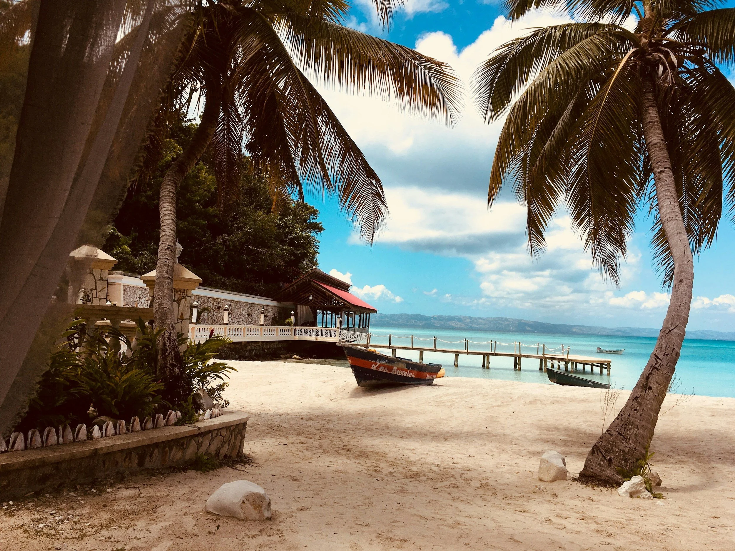 A tropical beach scene with palm trees, a small boat on the sand, and a dock leading out into clear blue water, with a partly cloudy sky.