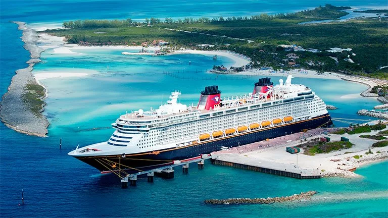 A large Disney Cruise Line ship docked at a pier in a tropical setting with clear blue water, sandy beaches, and lush green islands in the background.