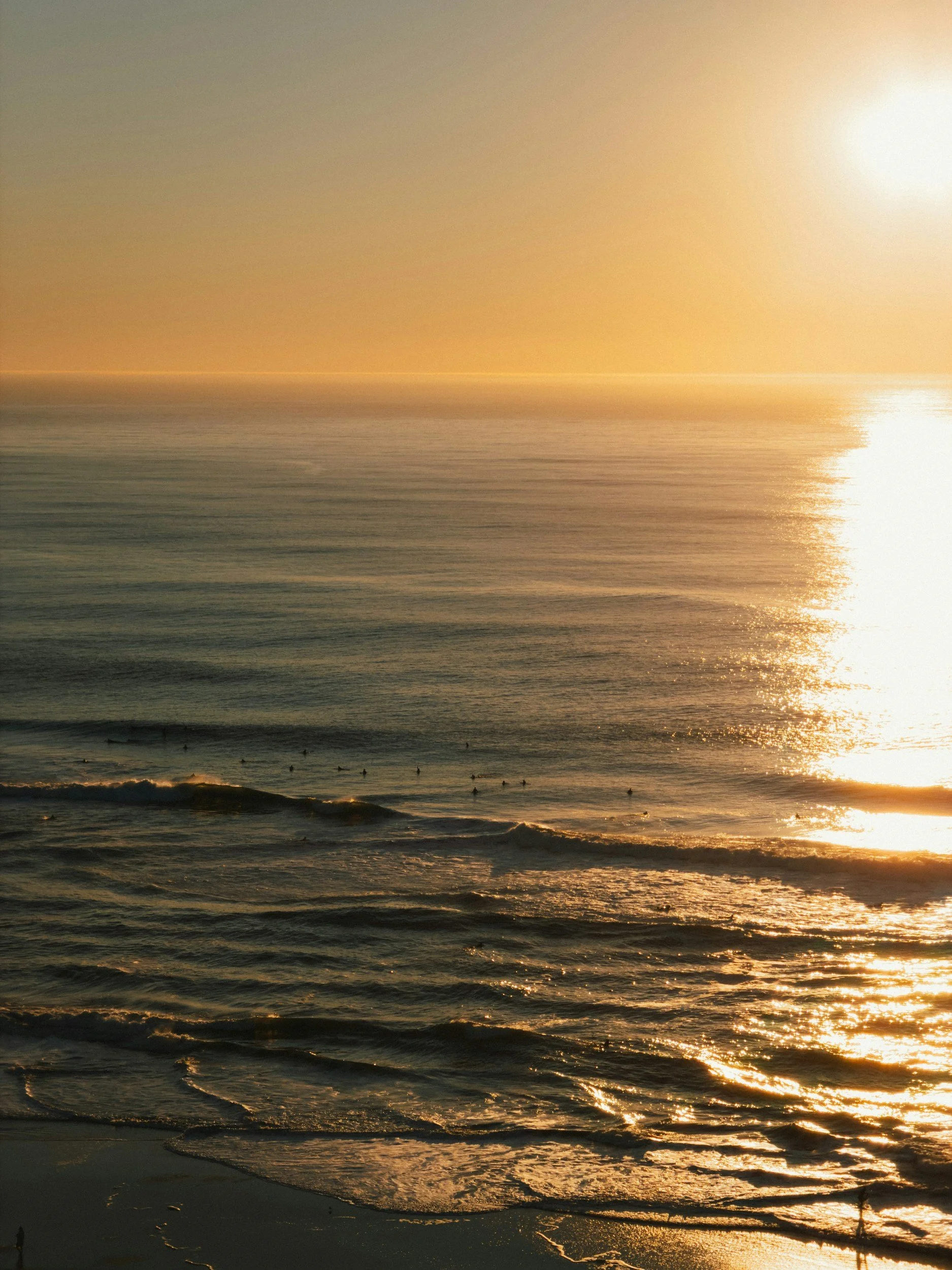 Sunset over the ocean with a clear sky, gentle waves, and surfers visible in the water.