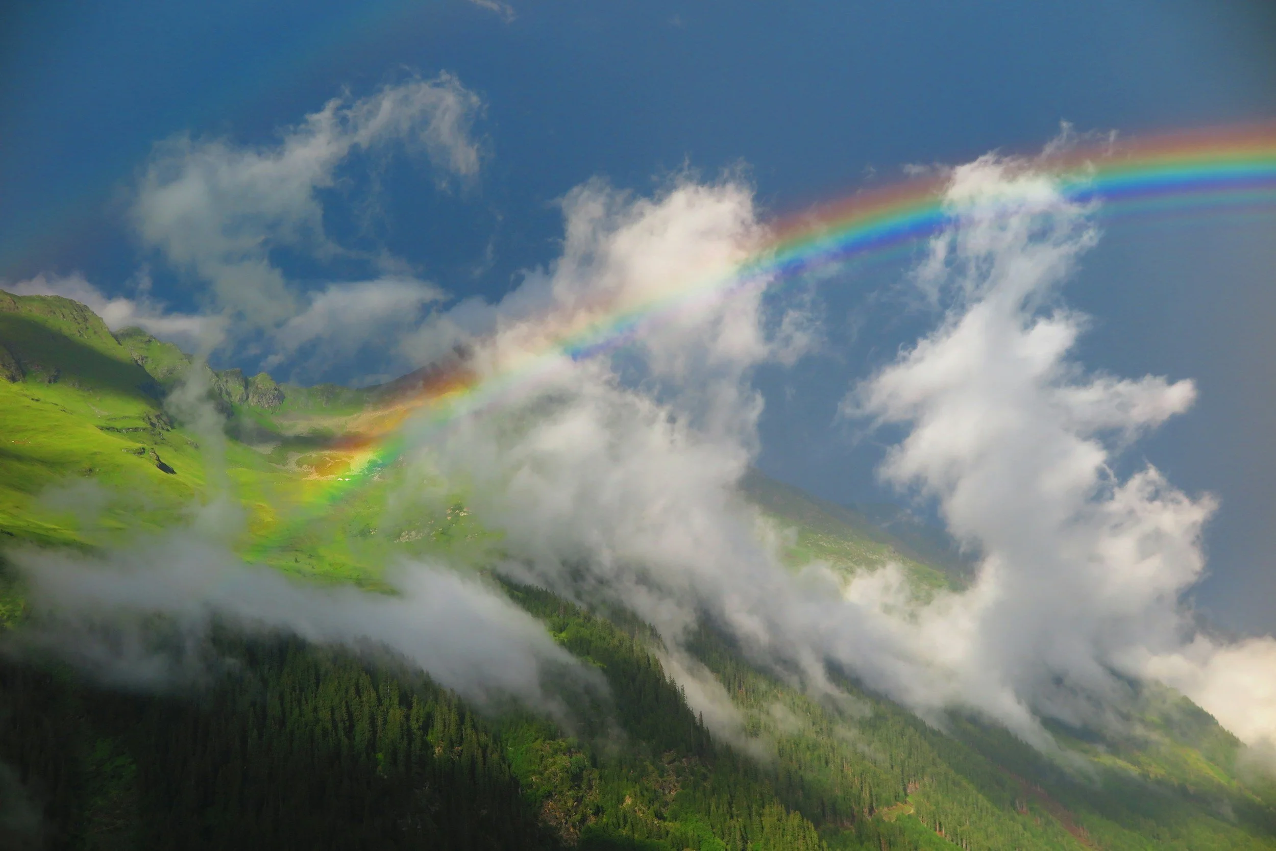 A rainbow arcs across a blue sky above green mountains and a forested valley, with clouds and mist in the air.