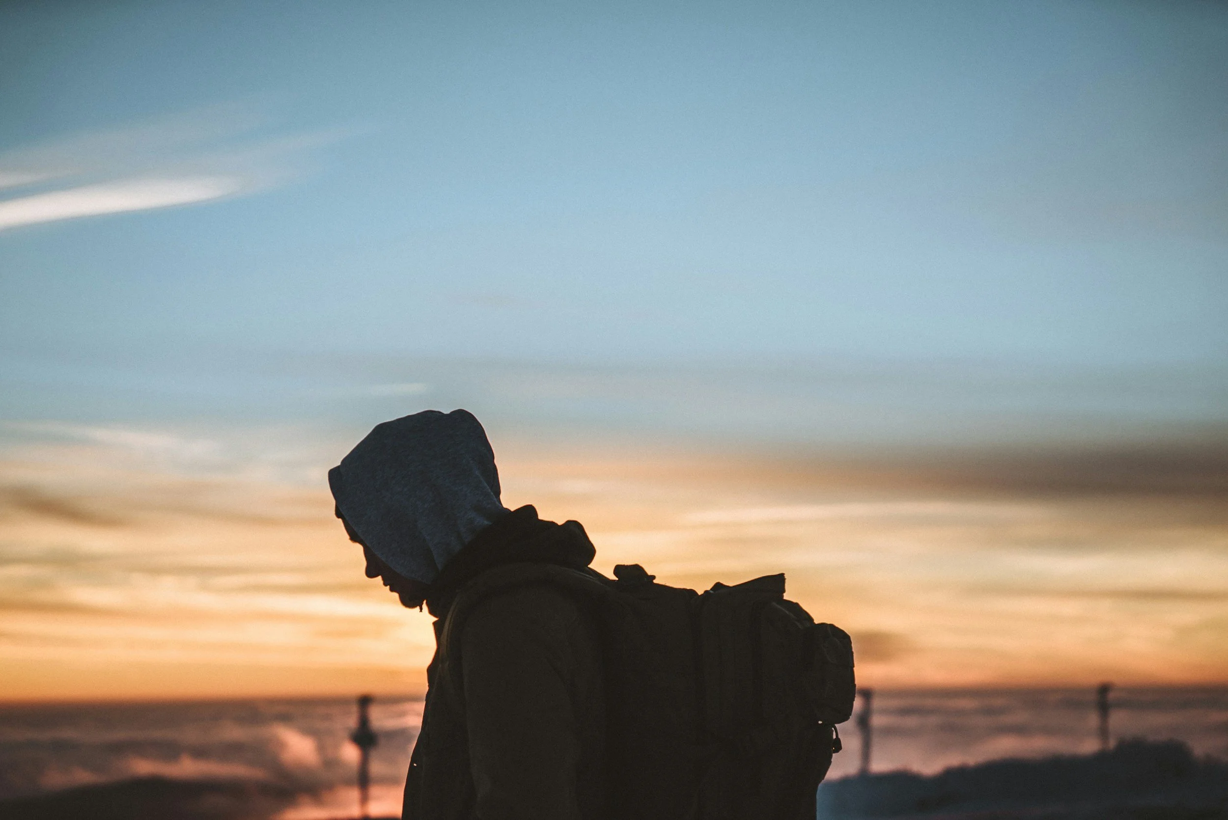 Person wearing a hoodie standing outdoors at sunset near a beach or coastal area.