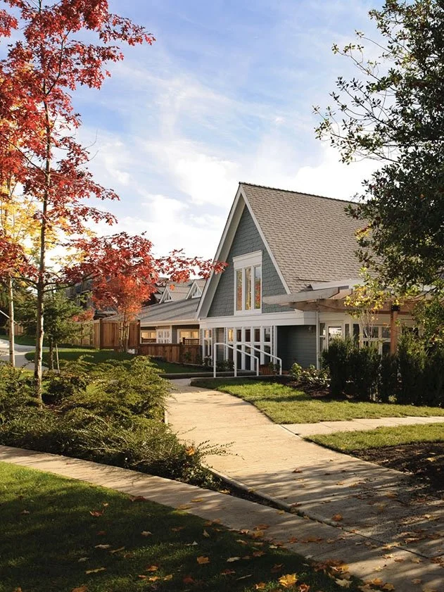 A suburban neighborhood with a house featuring a steep roof, a large front yard, a concrete walkway, and autumn trees.
