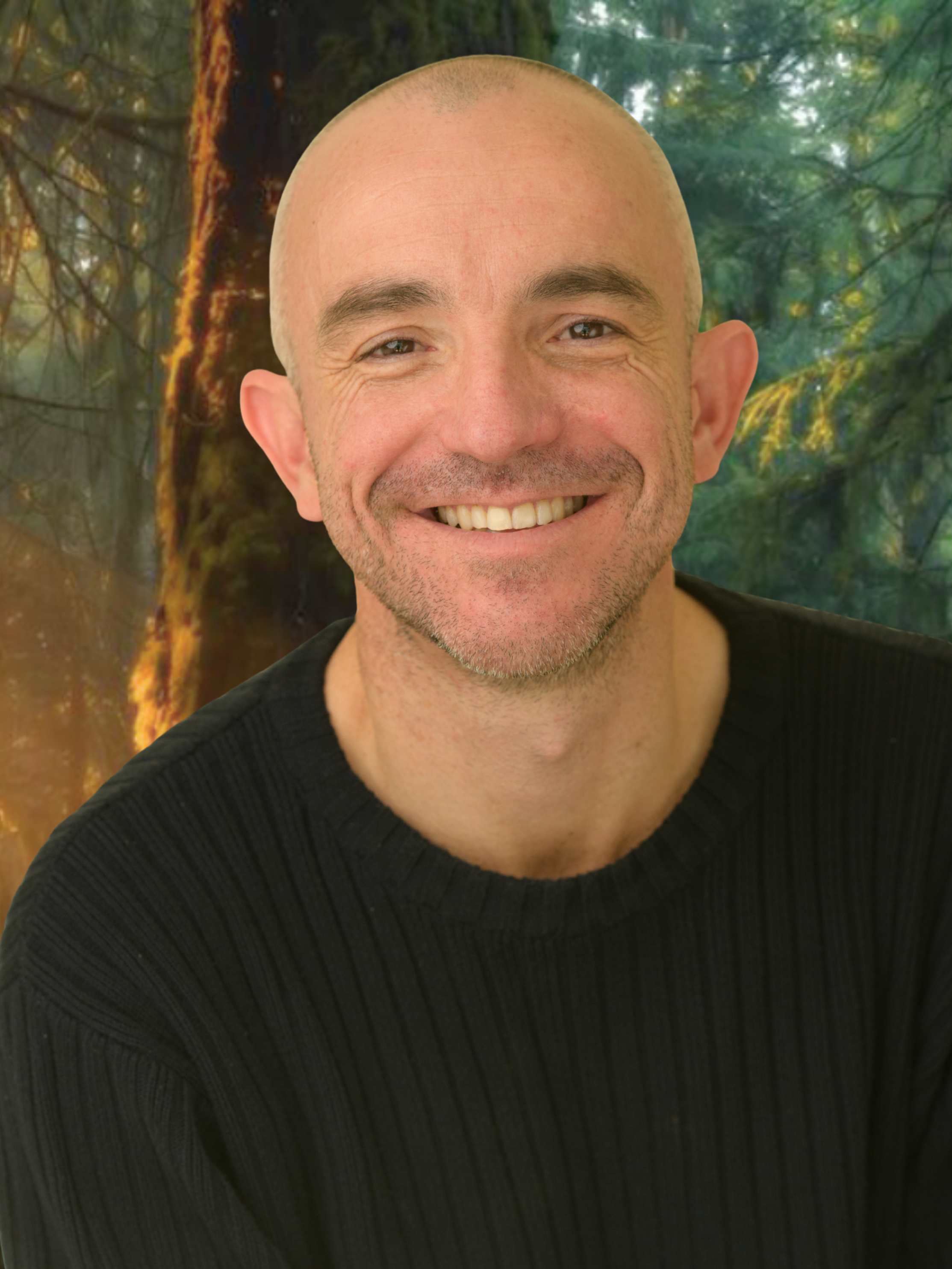 Close-up of a smiling, bald man with a light complexion, wearing a black ribbed shirt, with a blurred green forest background.