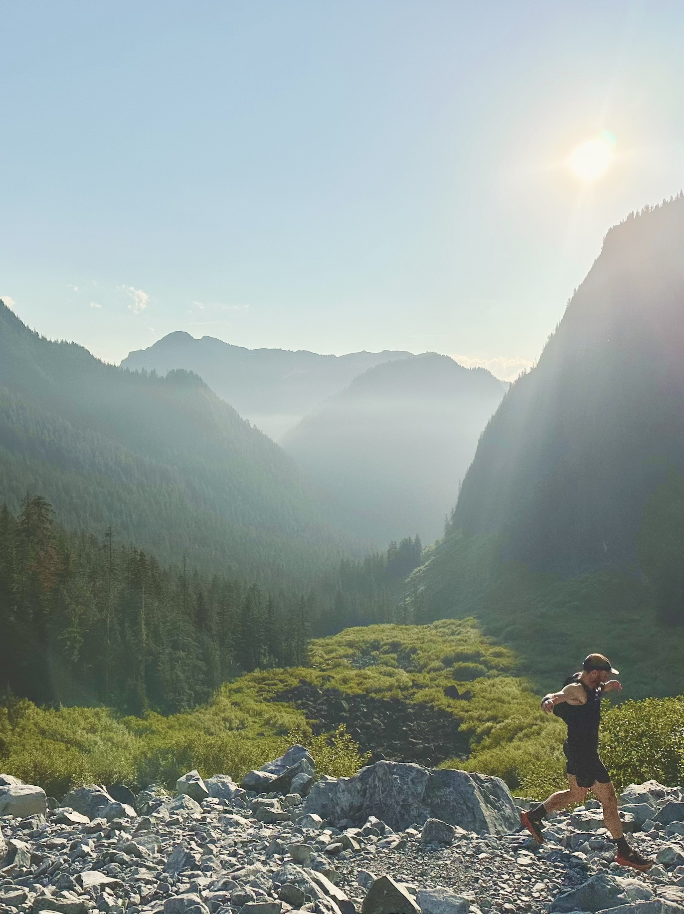 A person running on rocky terrain in a lush green valley surrounded by tall mountains under a bright sunny sky.