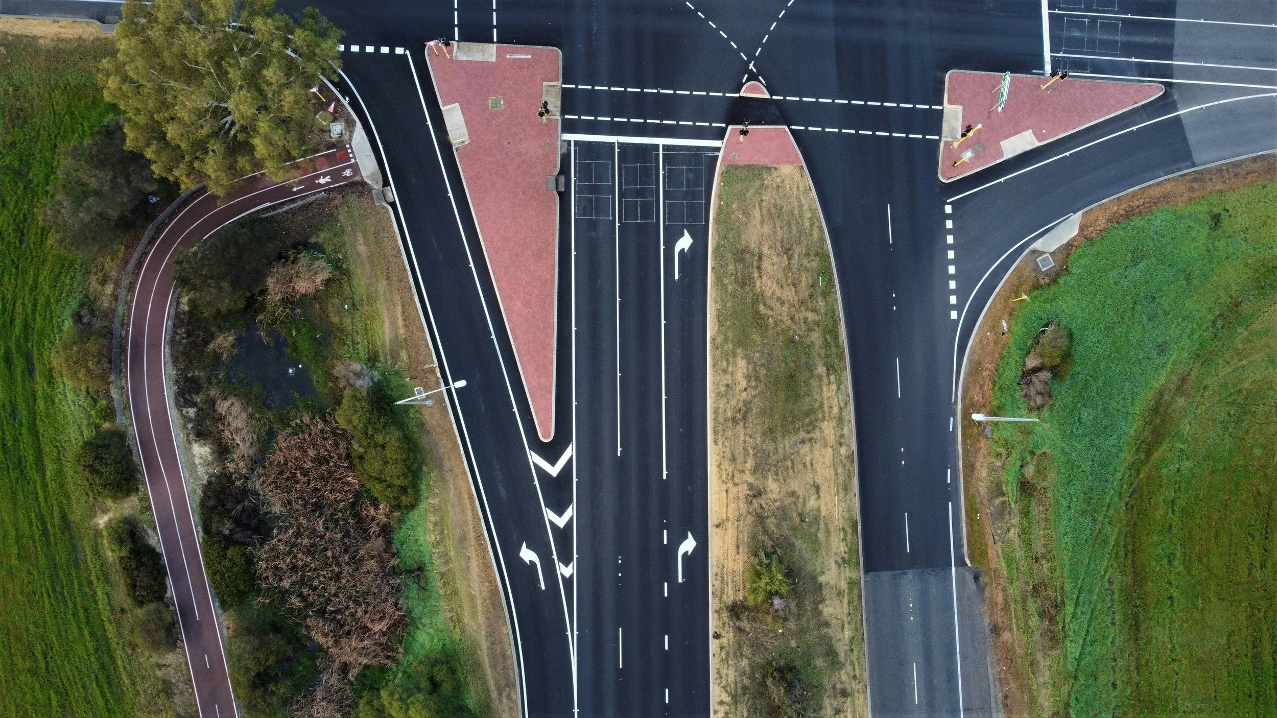 An aerial view of a newly paved intersection with multiple lanes, directional arrows, crosswalks, and traffic markings, surrounded by greenery, trees, and open land.