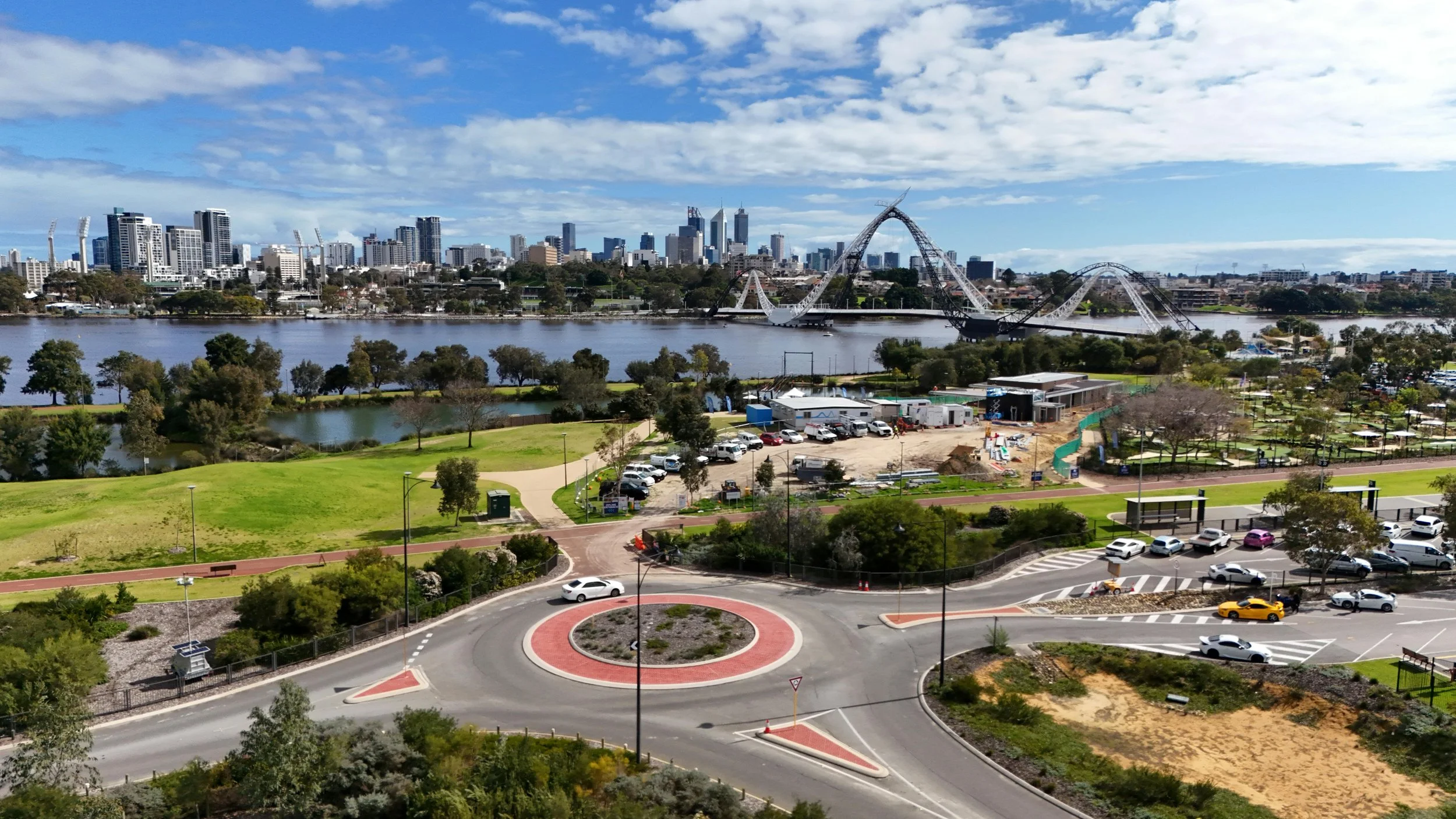 City skyline with modern buildings across a body of water, featuring a large roller coaster, green park, and parking lot in the foreground.