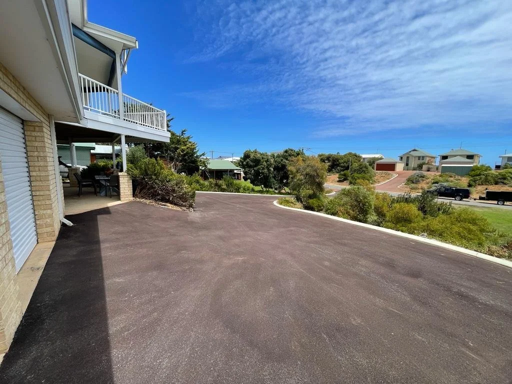 A large asphalt driveway in a suburban neighborhood, with houses on a hill and a blue sky with some clouds.