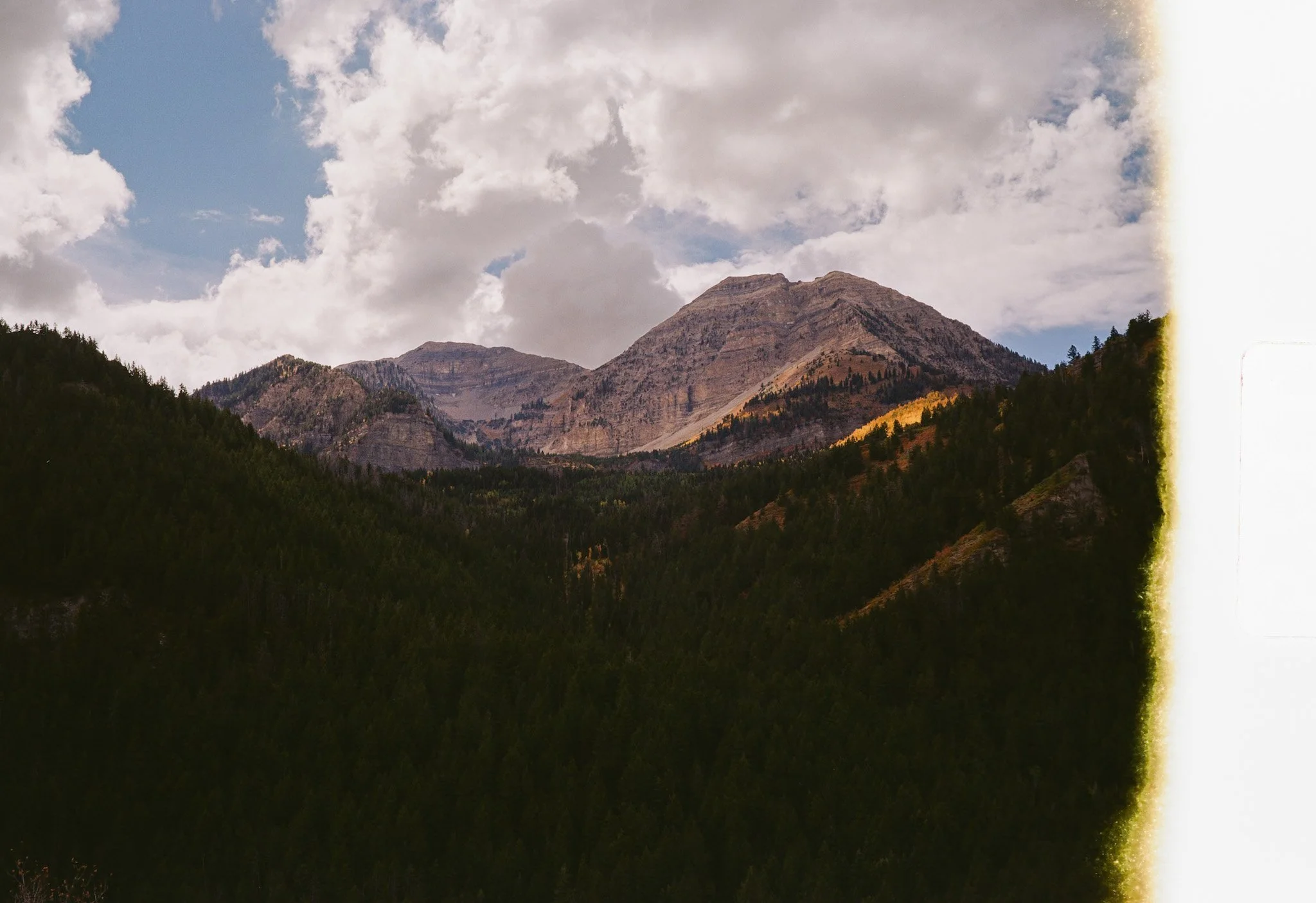 Mountain landscape with forested slopes, rocky peaks, and a partly cloudy sky.