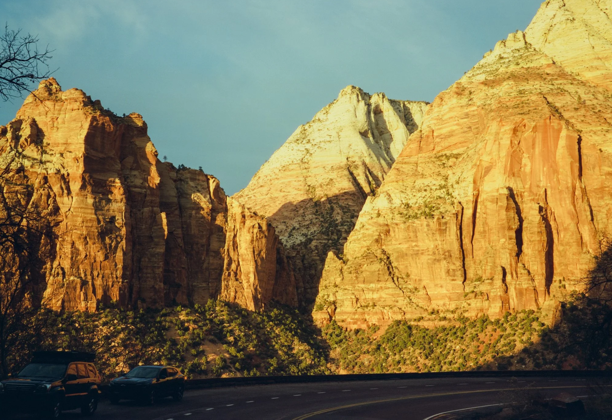 Sunlit mountains with red and white rock formations, green shrubbery at the base, and a paved road with parked cars in the foreground.