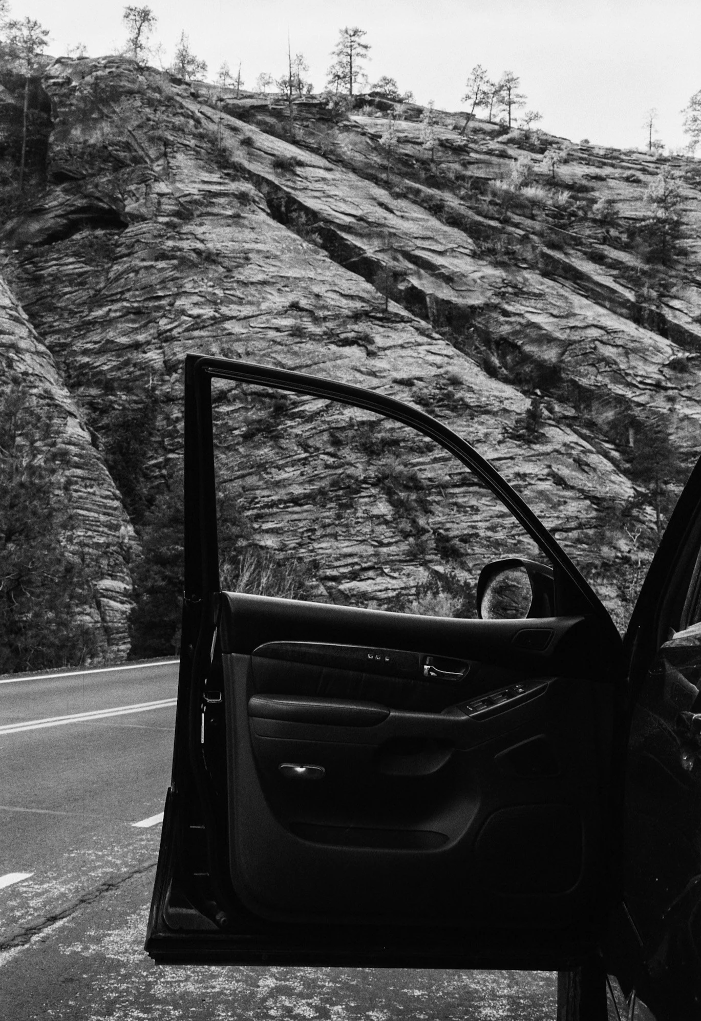 A view of a rocky mountain landscape through the open driver-side door of a car, parked on a road.