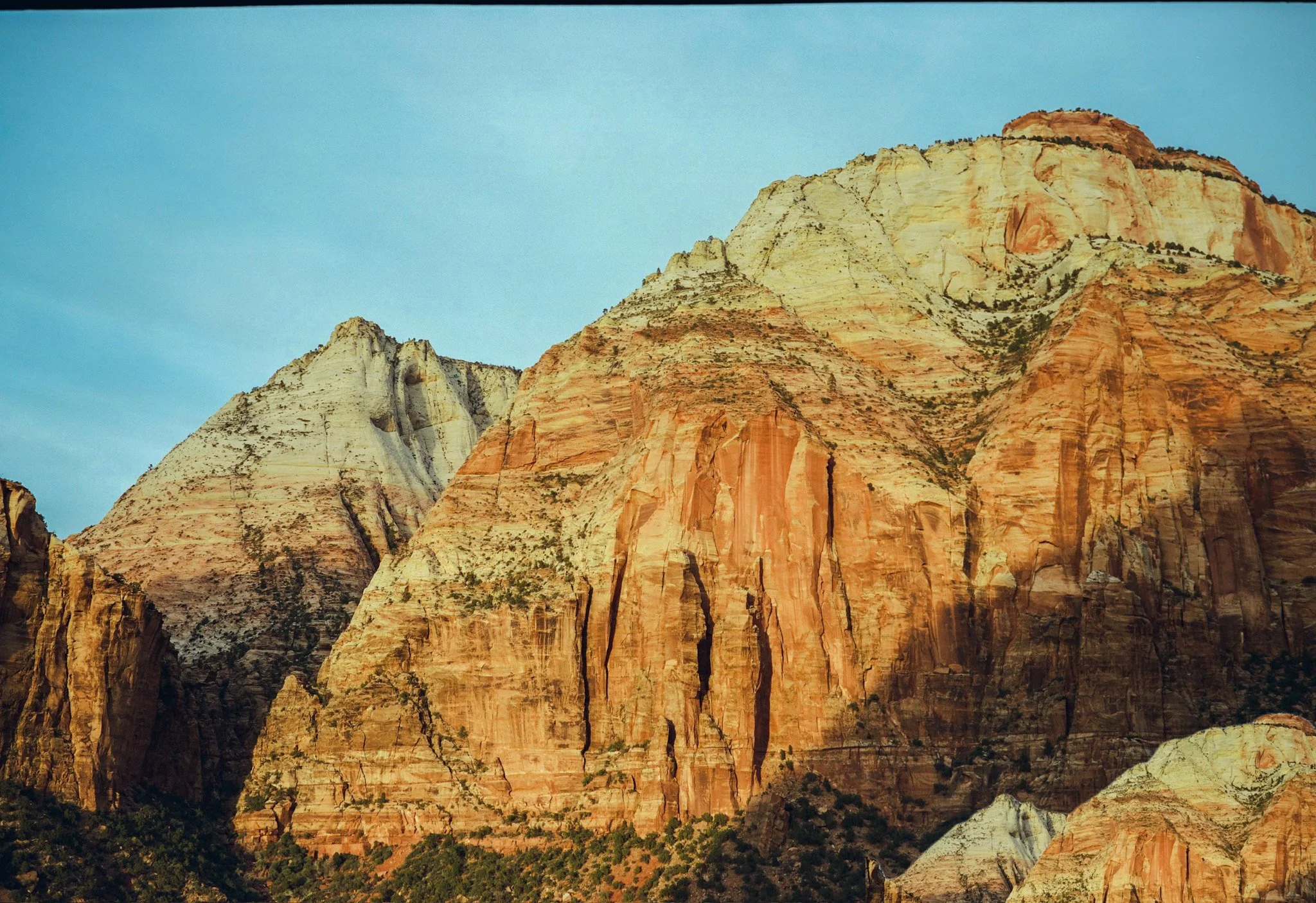 Colorful mountain cliffs with layered rock formations in shades of red, orange, and white, with some green vegetation at the base under a clear blue sky.