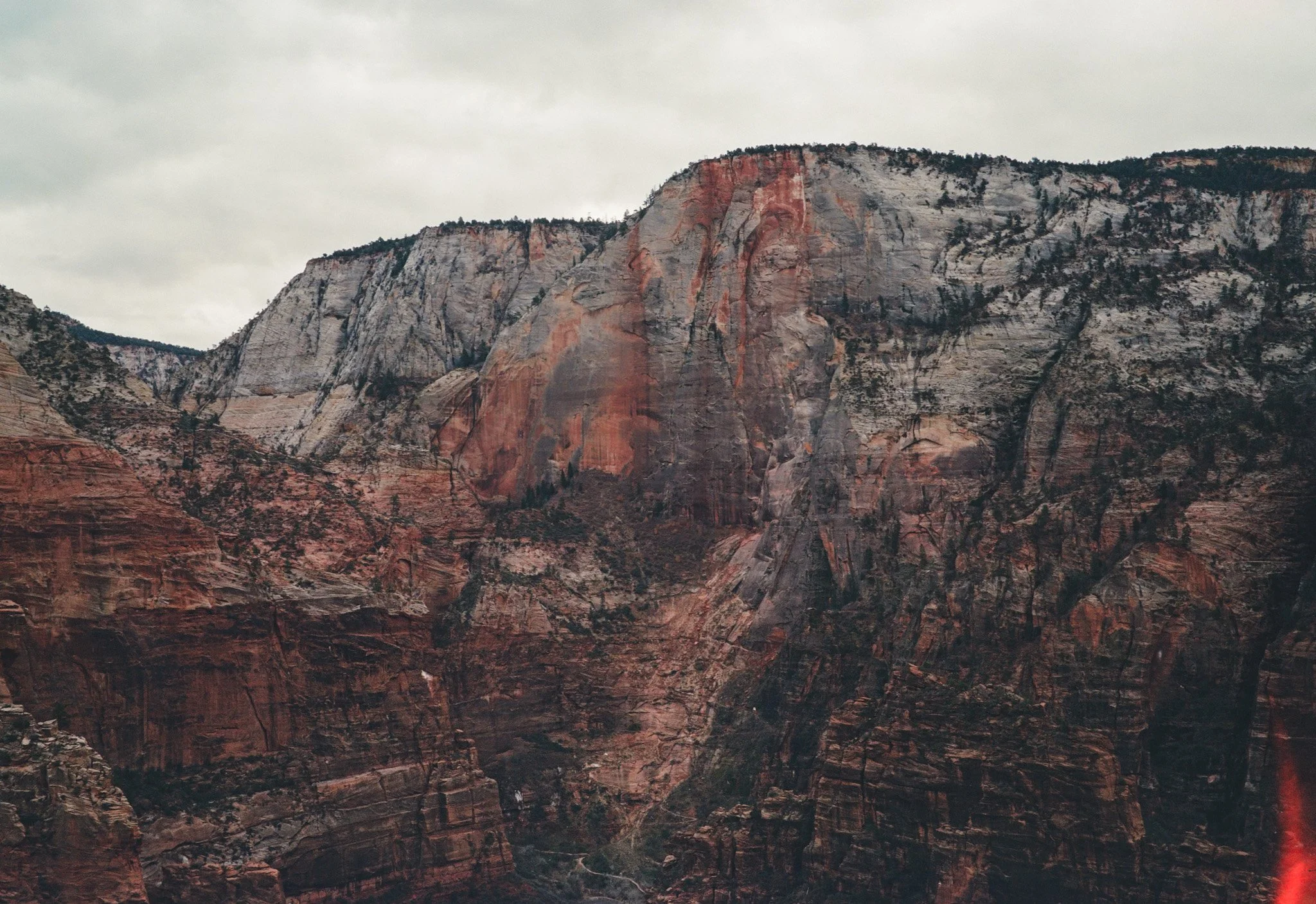 A rugged canyon landscape with steep, colorful rock cliffs and sparse vegetation under a cloudy sky.