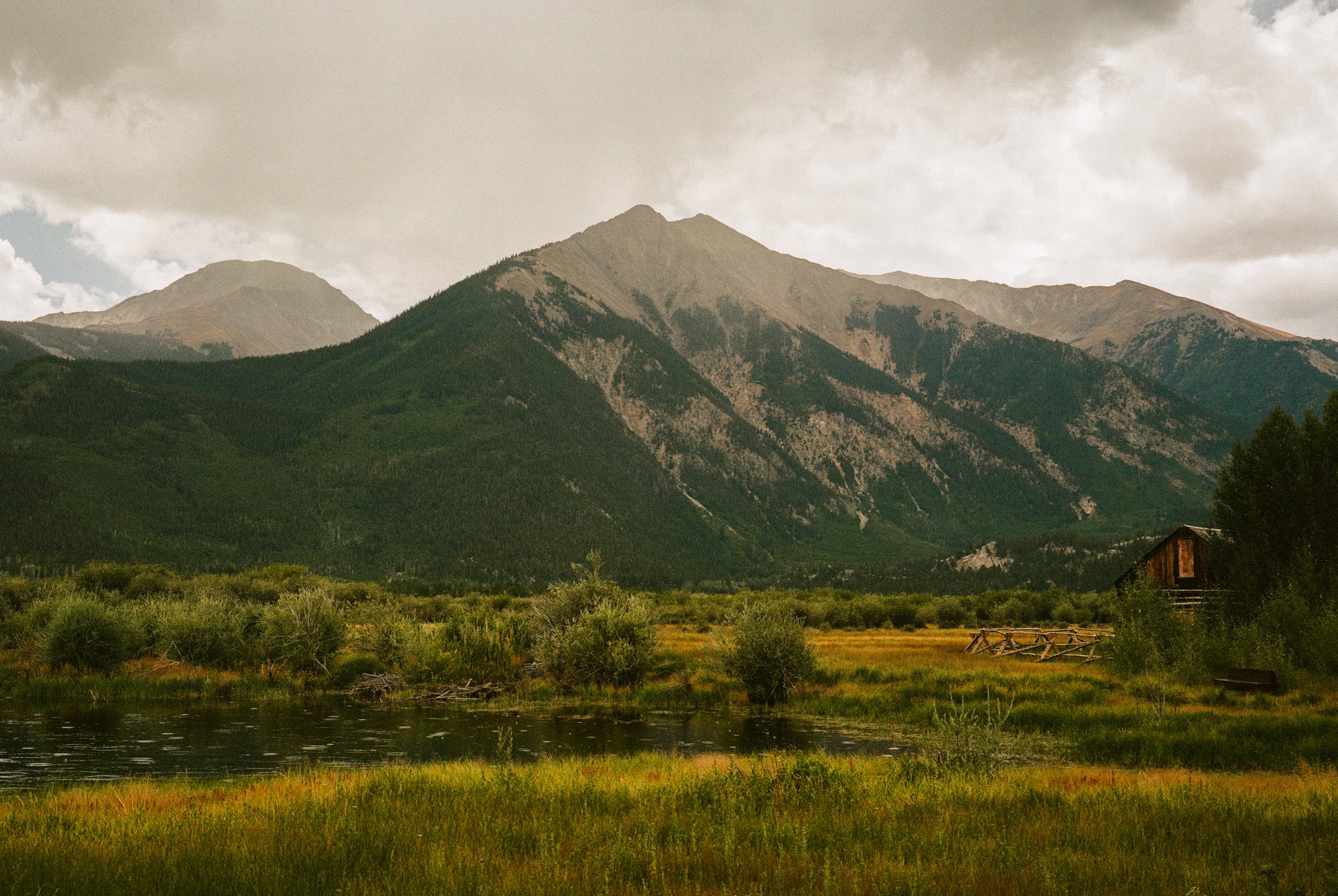 A landscape of green mountains under a cloudy sky, with a small pond, trees, and a rustic wooden barn in the foreground.