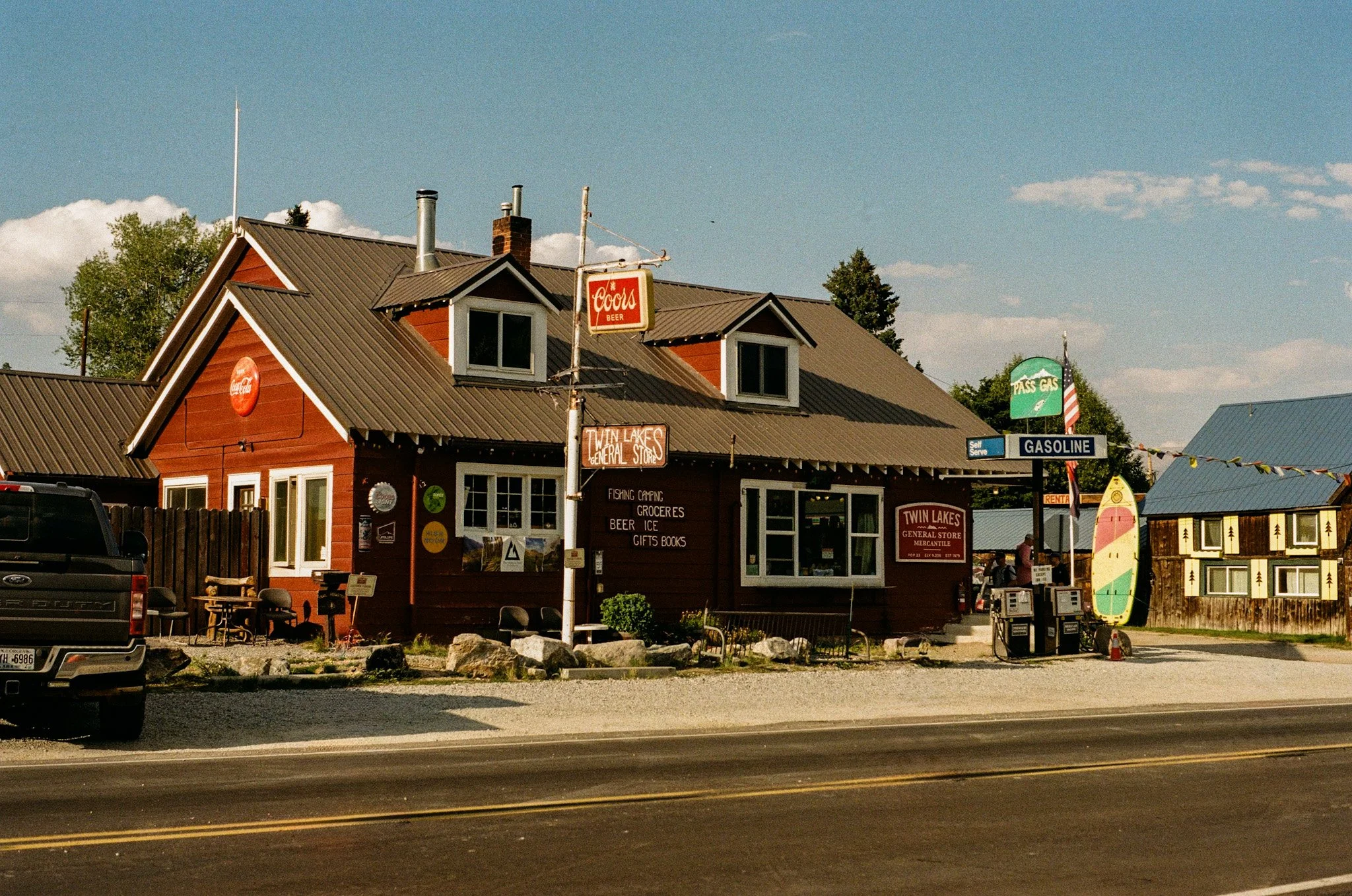 A red wooden general store called Twin Lakes, with signs indicating it sells fishing supplies, groceries, beer, ice, gifts, and books. There is a surfboard sign out front, and a nearby gas station sign. The store is located on a gravel lot next to a 