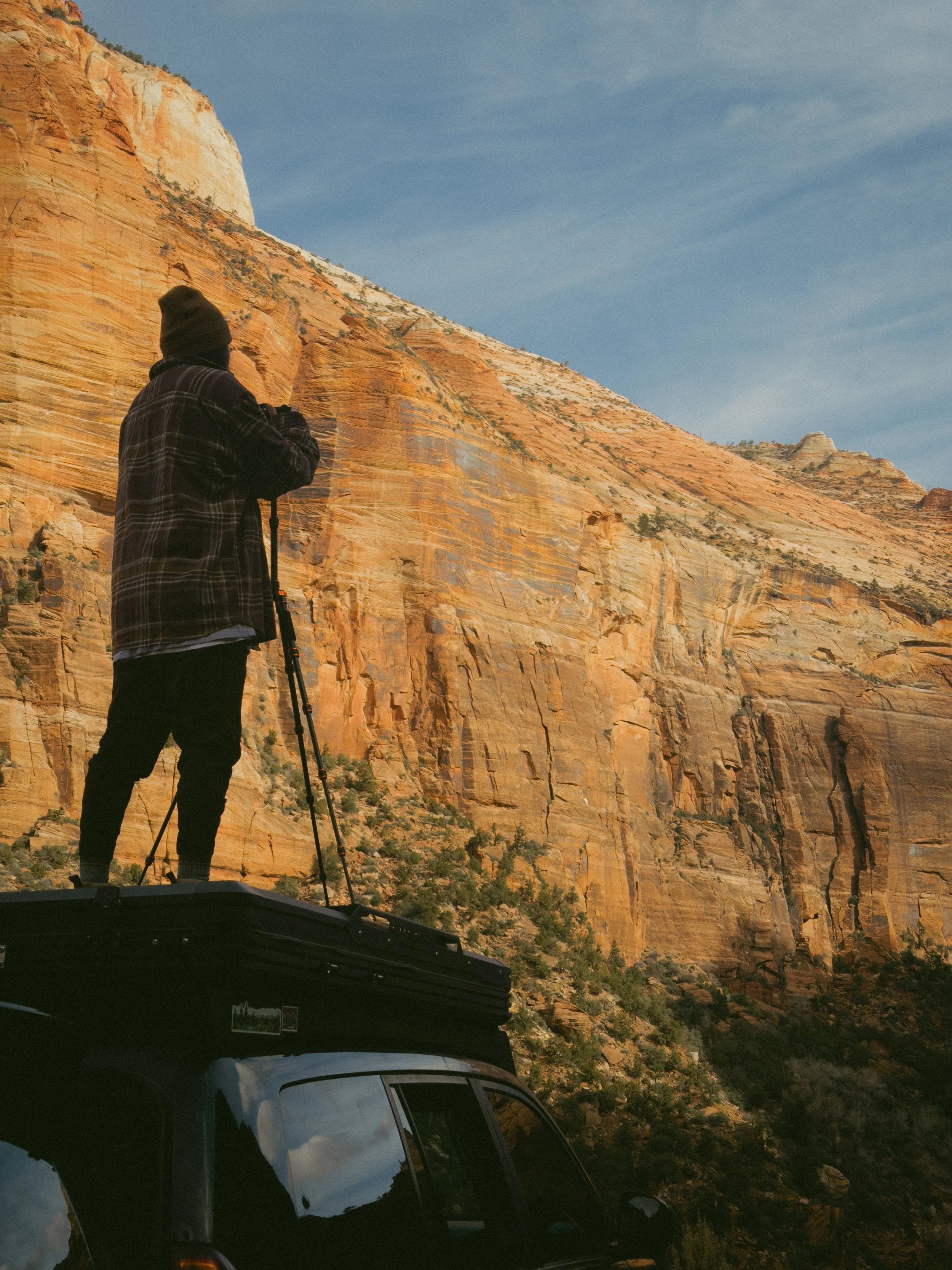 Person standing on top of a vehicle, using a tripod, taking a photograph of a large, orange rock formation under a blue sky.