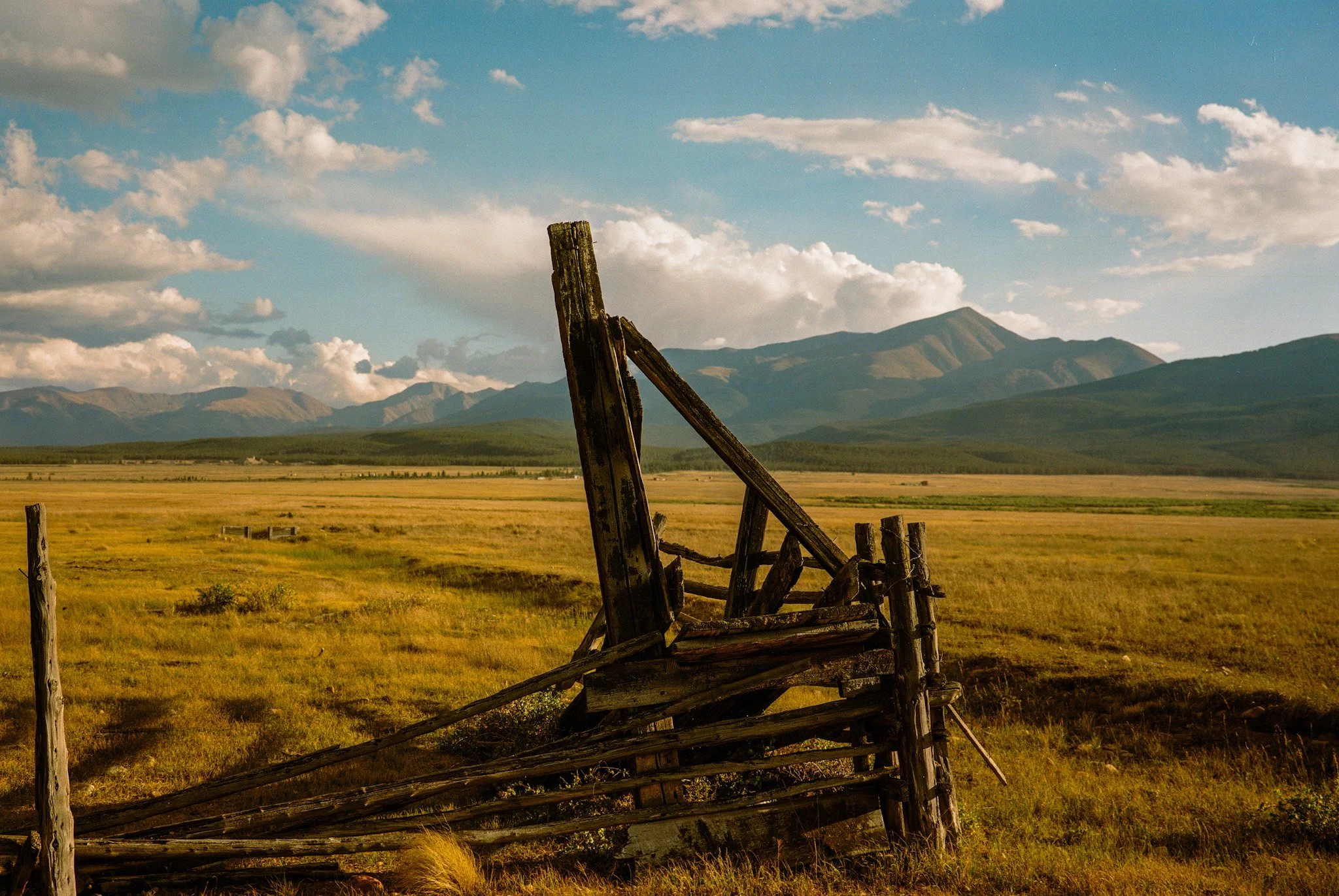 An old, weathered wooden fence standing in a vast grassy field with mountains in the background, under a partly cloudy sky.