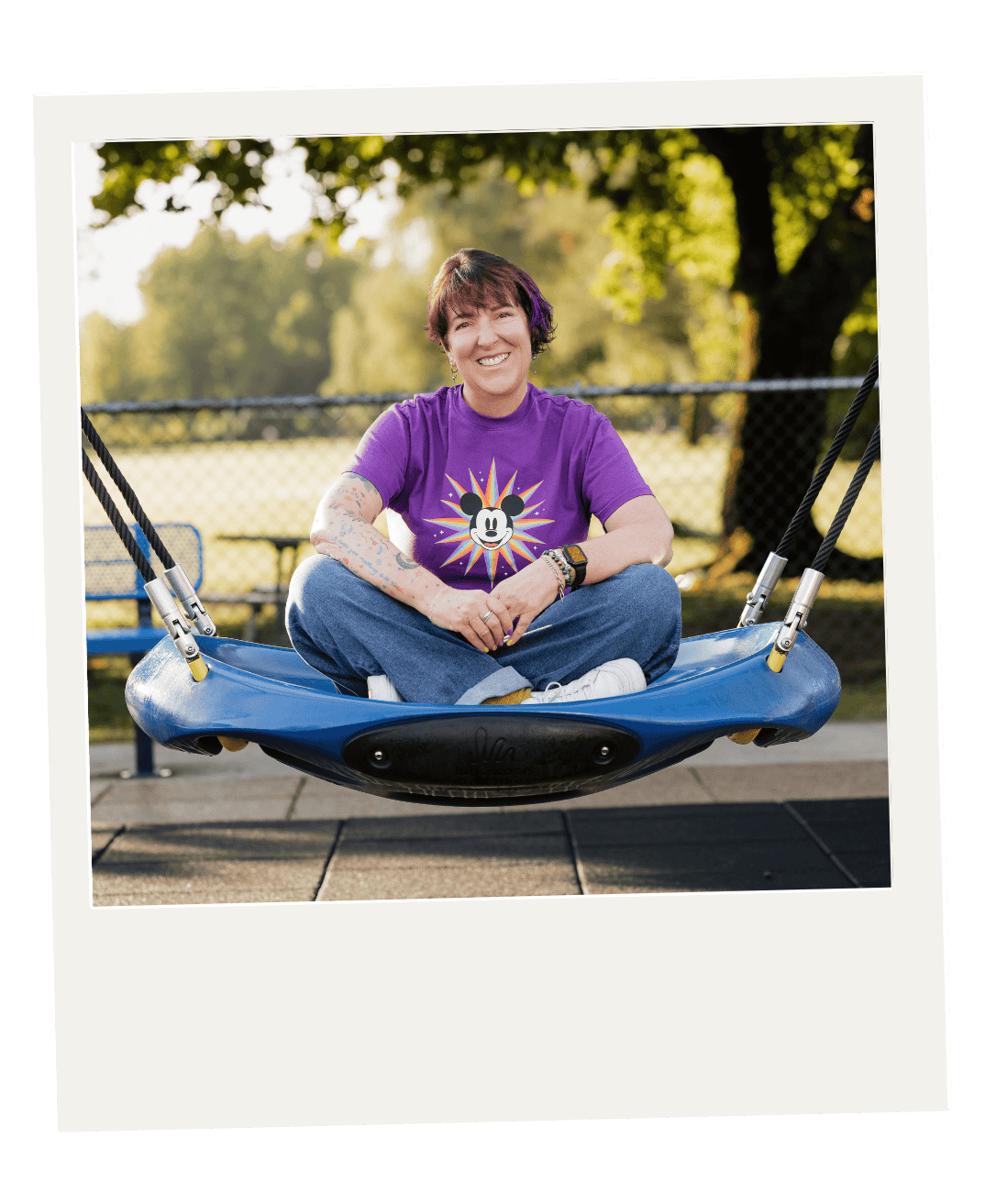 A smiling woman with short brown hair sitting cross-legged on a blue swing at a park, wearing a purple t-shirt with Mickey Mouse and colorful starburst design, and jeans, with trees in the background.