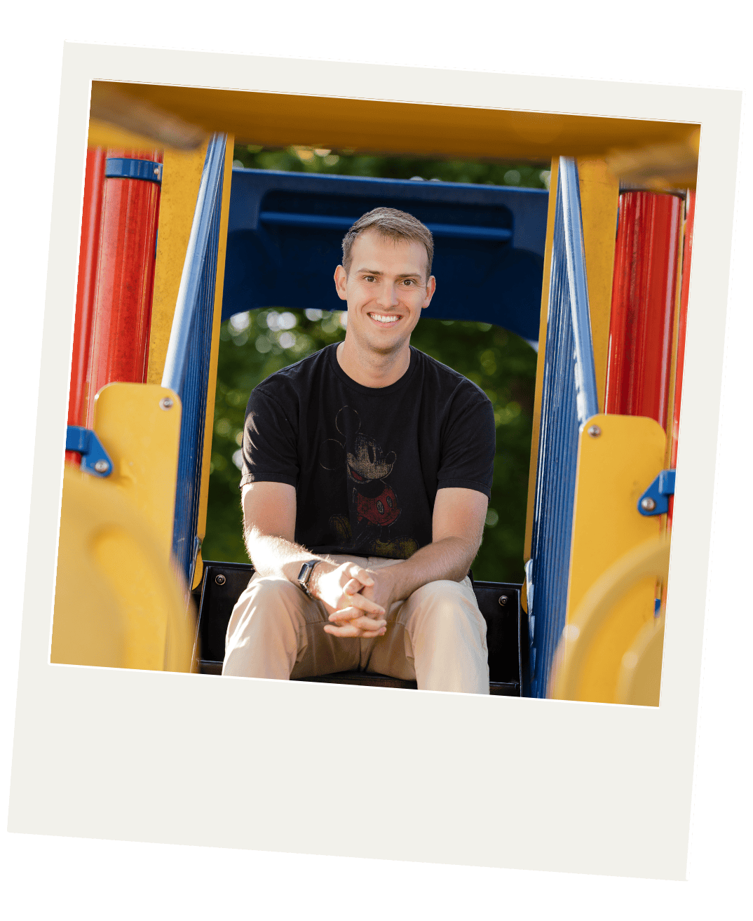 A smiling man sitting on a slide in a playground, wearing a black t-shirt with Mickey Mouse, beige pants, and a wristwatch, with trees in the background.