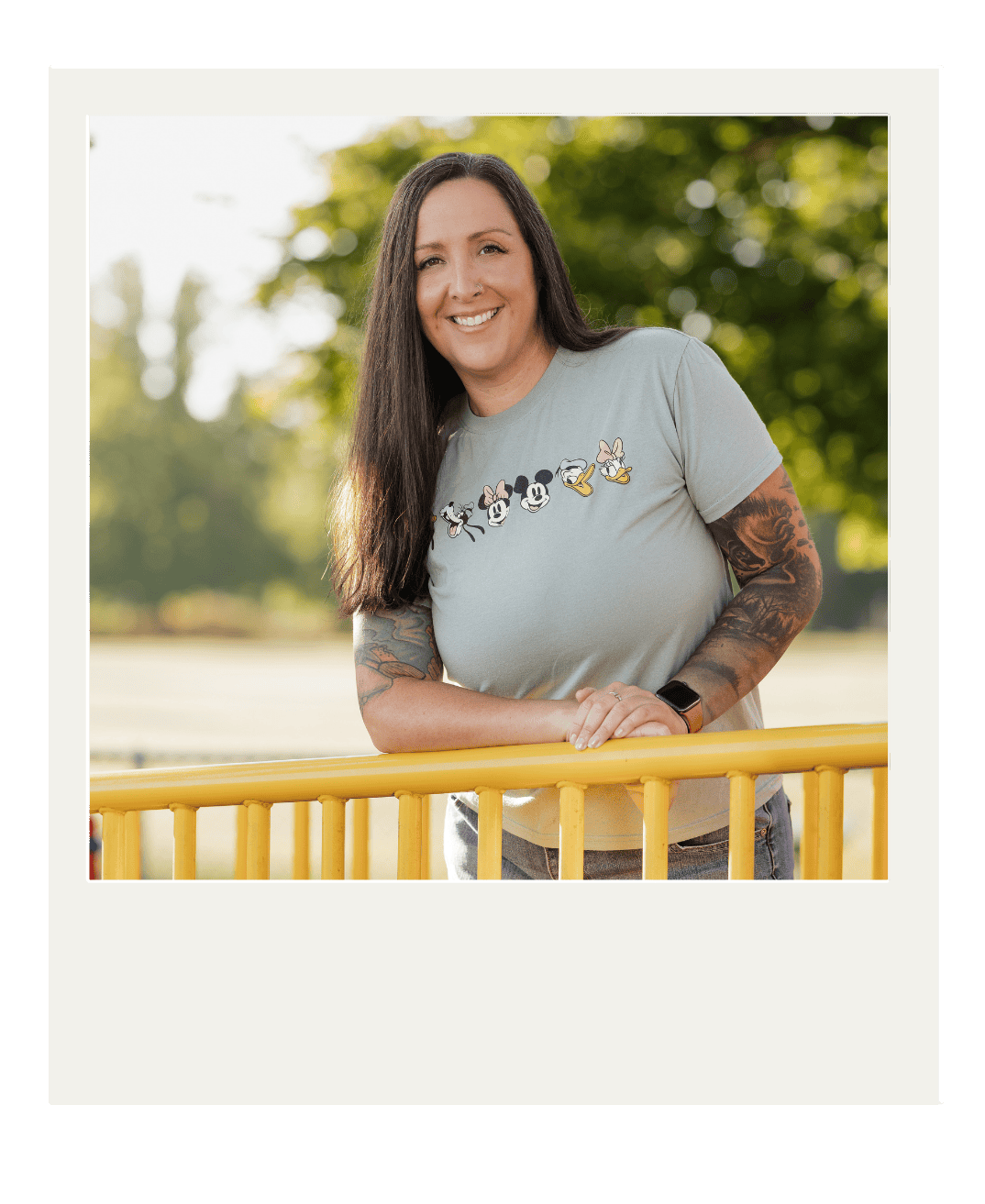 A woman with long dark hair and tattoos, wearing a gray T-shirt with Disney character illustrations, leaning on a yellow railing outdoors with green trees in the background, smiling at the camera. Rosa is our office coordinator. 