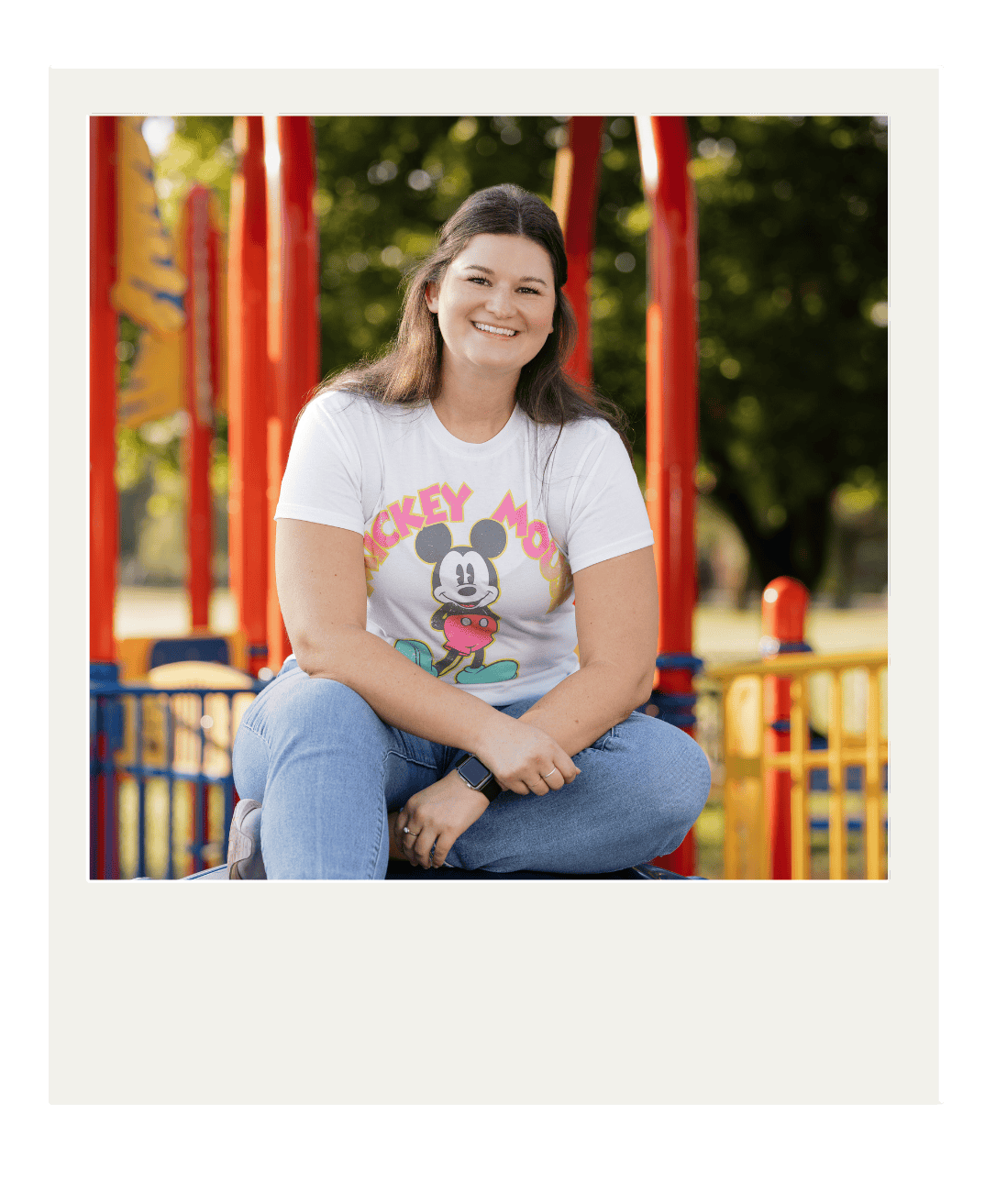 Woman with long brown hair smiling, wearing a Mickey Mouse t-shirt and jeans, sitting cross-legged on a playground structure in a park.