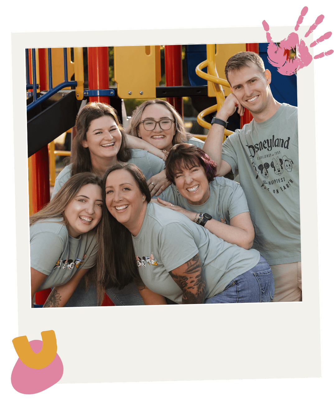 A group of Pediatric clinic staff posing together outdoors in front of playground equipment, wearing matching gray t-shirts, during daytime.
