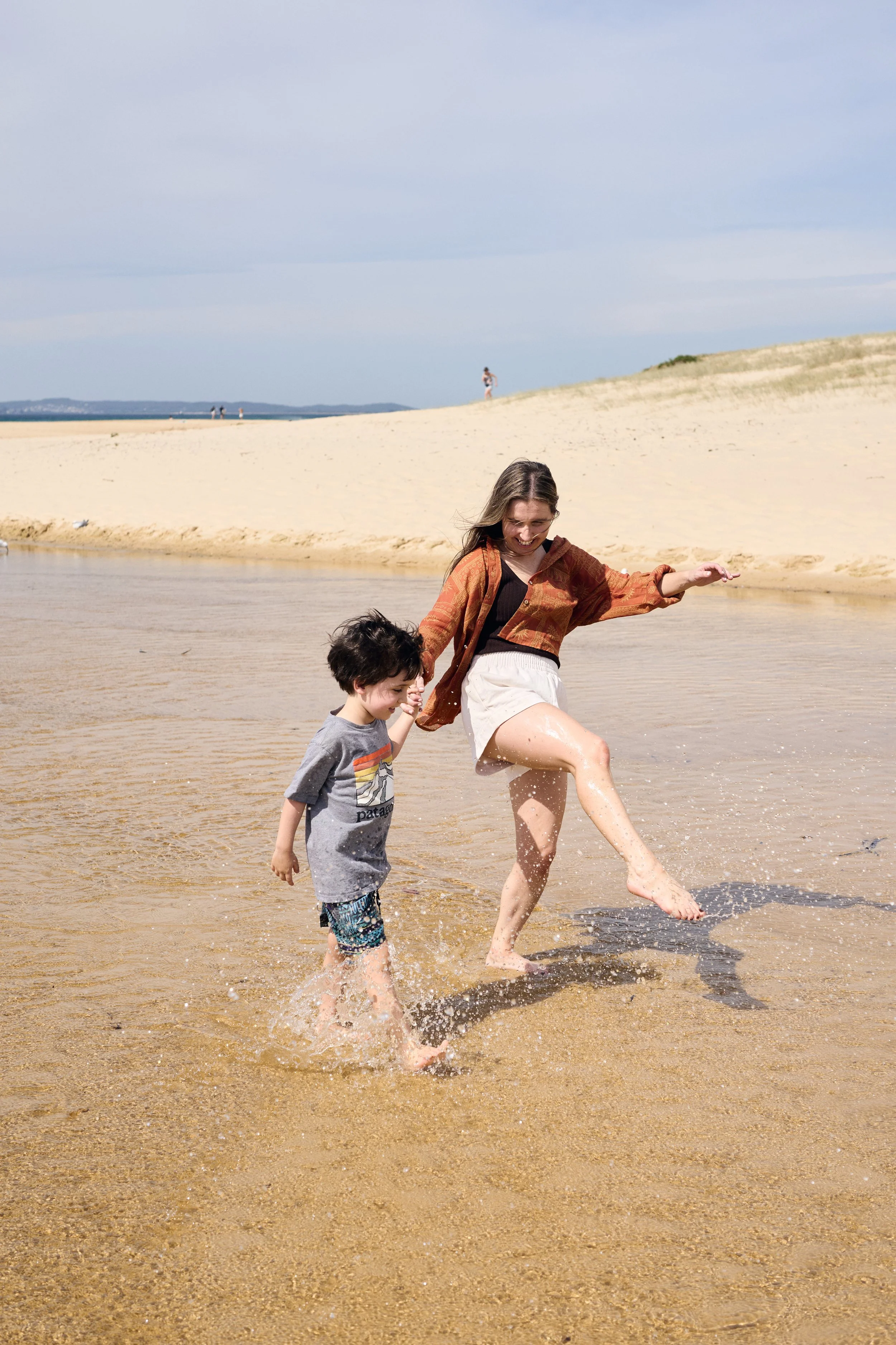 A woman and young boy playing and splashing water at a sandy beach, with dunes and a partly cloudy sky in the background.