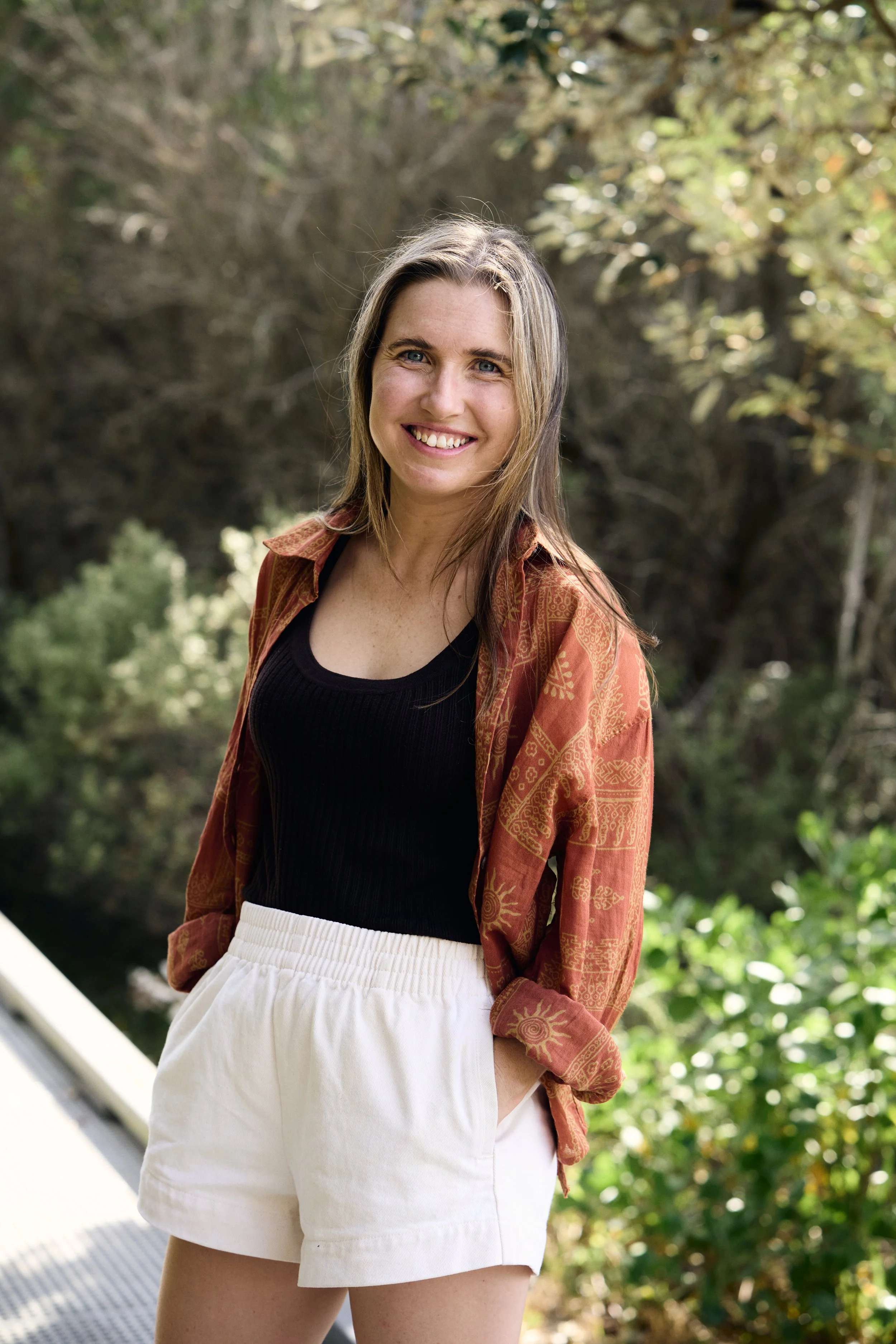 A young woman smiling outdoors in a natural setting with trees and greenery, wearing a black top, patterned jacket, and white shorts.