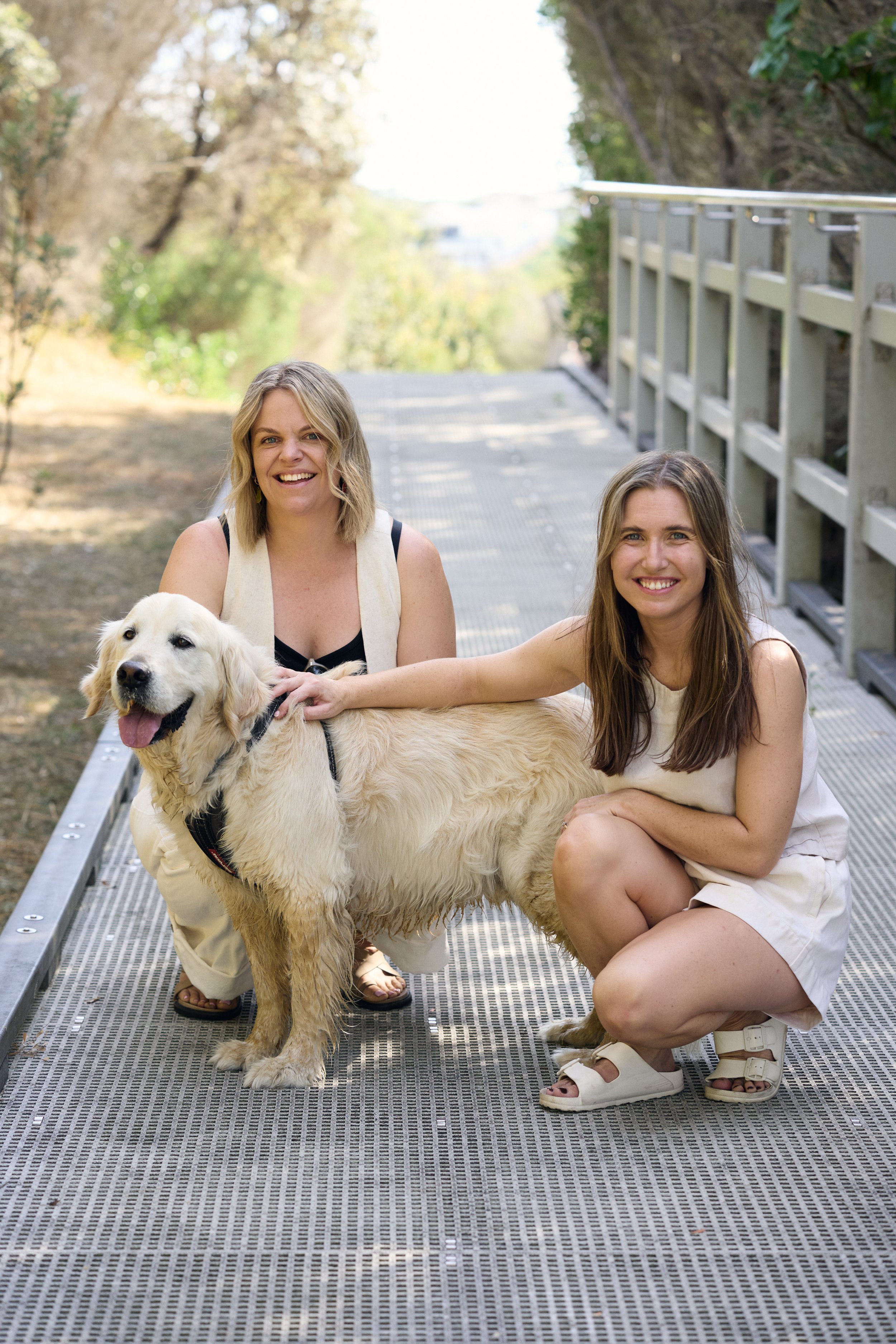 Two women and a golden retriever dog on a metal bridge outdoors, smiling at the camera.