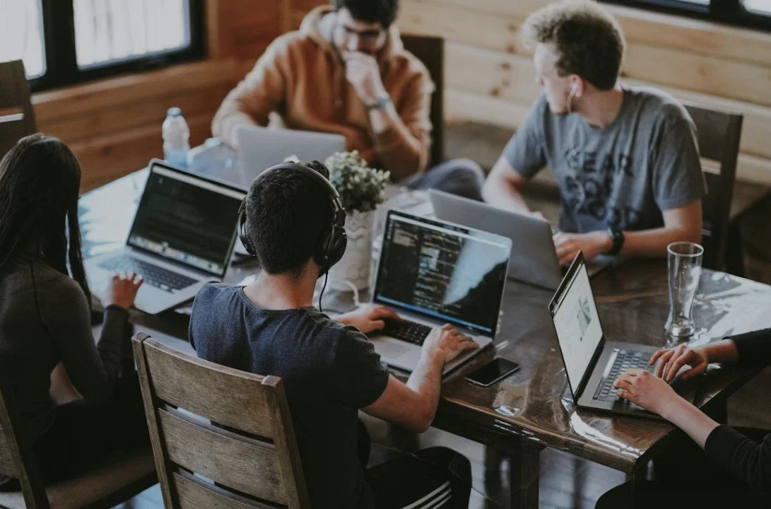 Group of five young adults working on laptops around a wooden table in a cozy room with wooden walls.