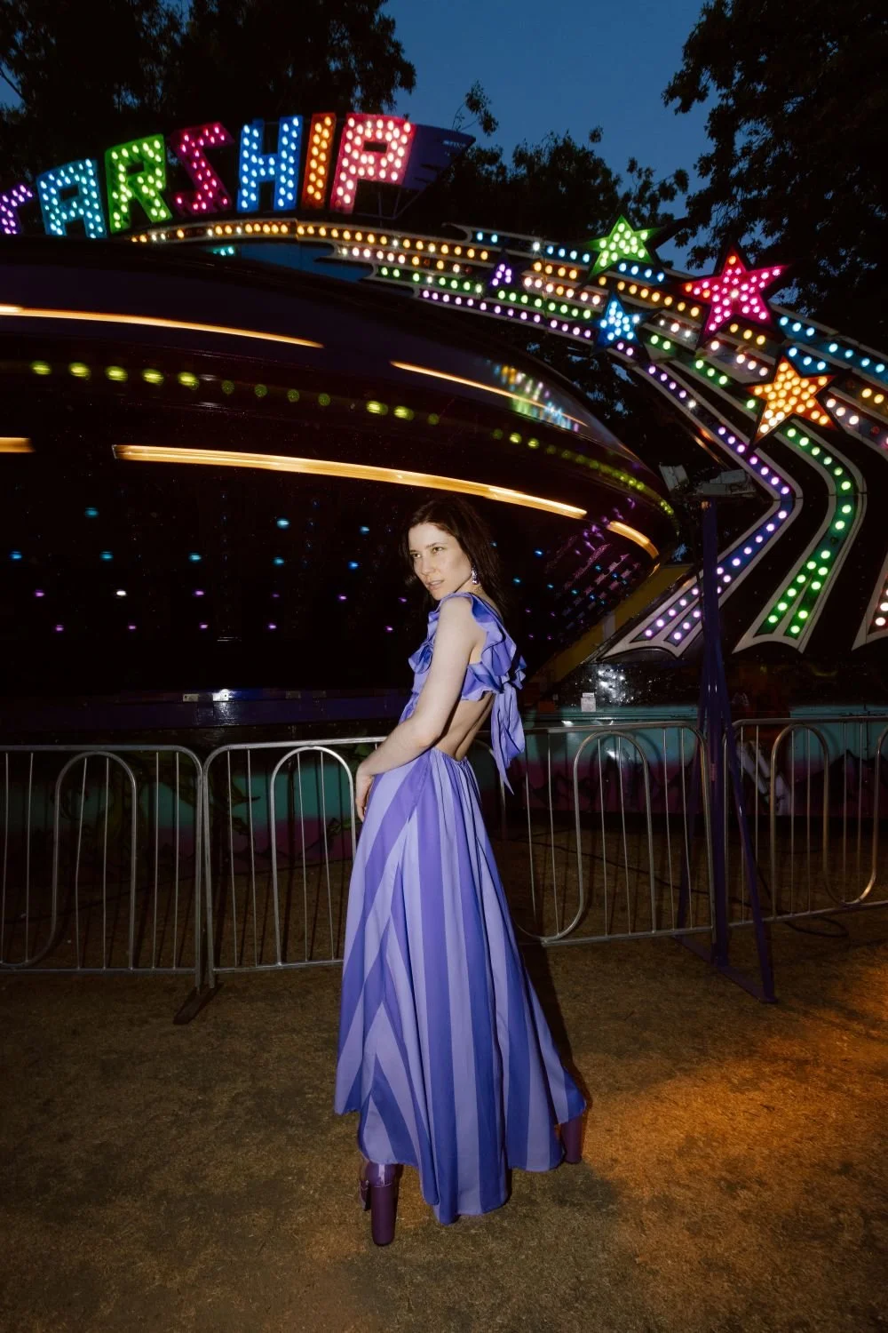 Krissy Eliot in a purple dress with ruffled shoulders and high heels stands in front of a carnival spaceship ride with bright, colorful lights at dusk