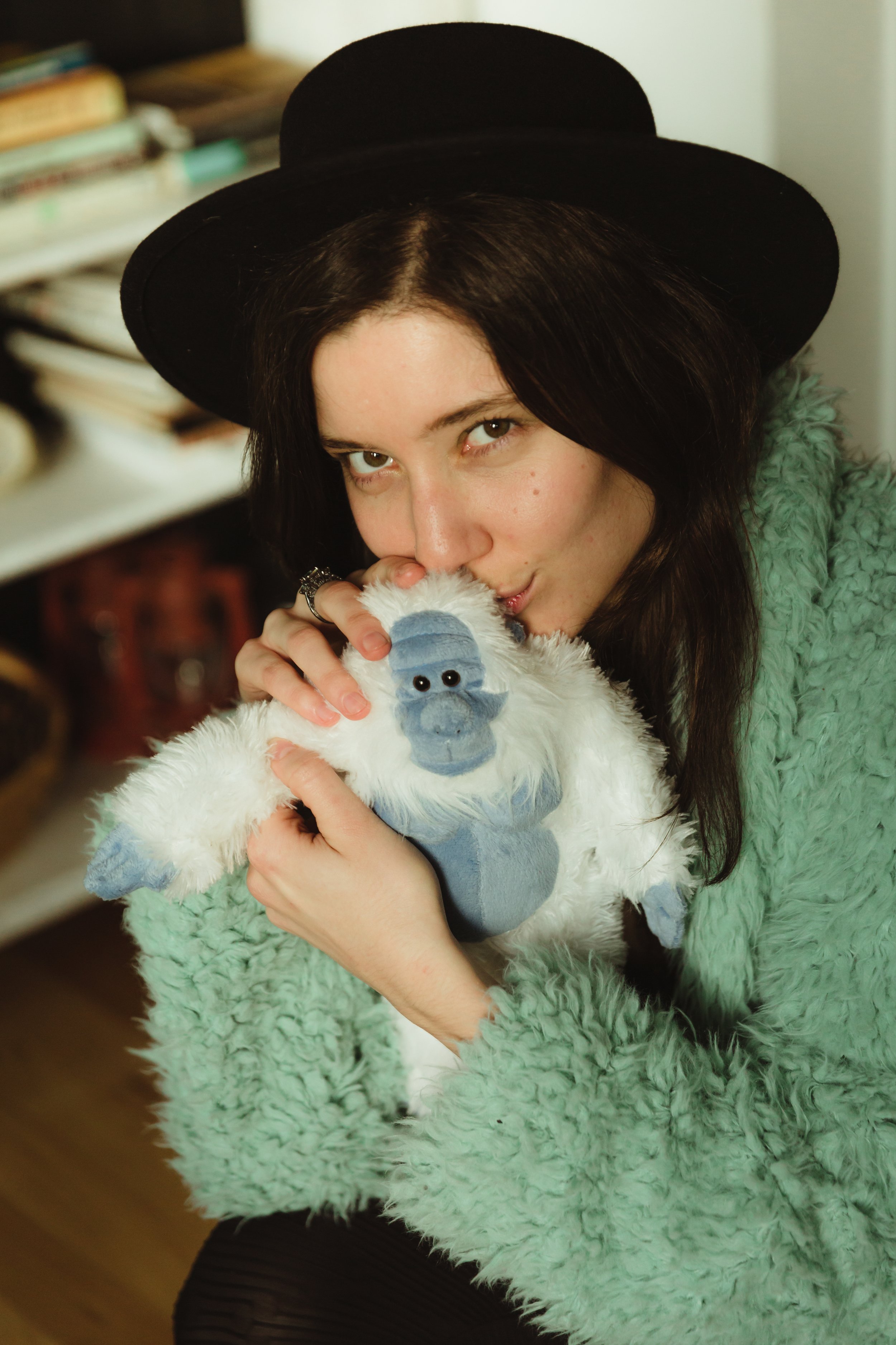 Krissy Eliot with dark hair, wearing a black hat and a mint green faux fur coat, is holding a plush toy of a white and blue yeti and kissing it