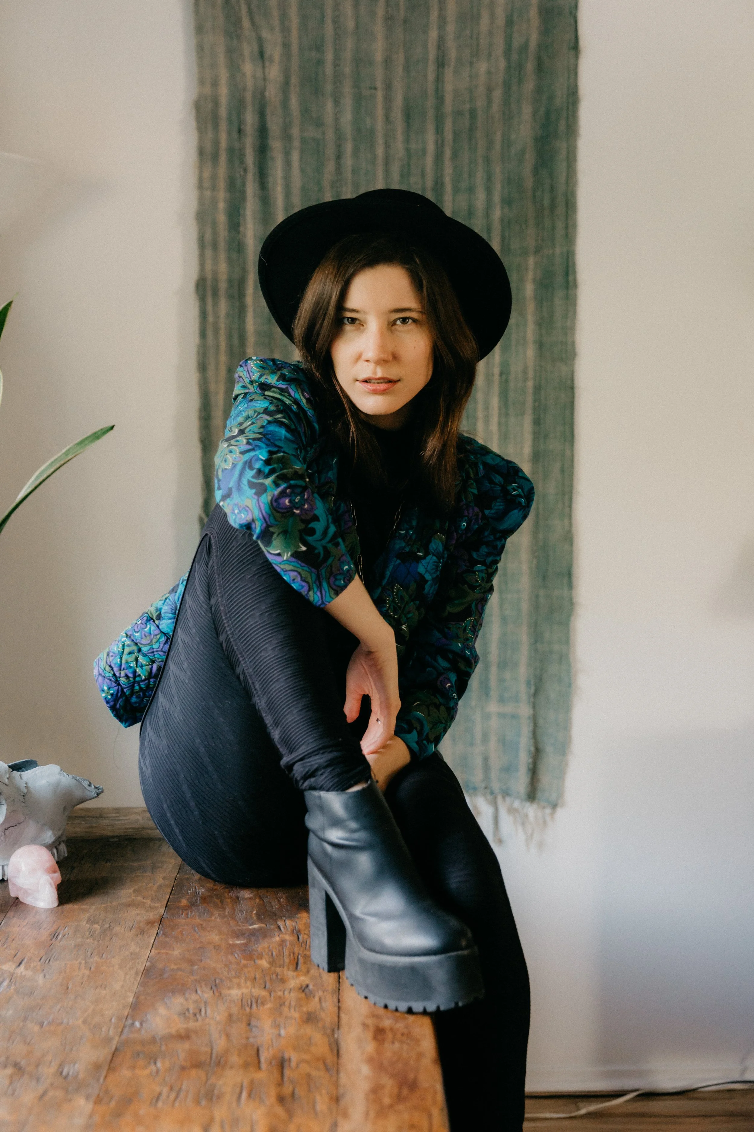 Krissy Eliot wearing a black hat, a colorful patterned jacket, black pants, and high-heeled boots, sitting on a wooden table in a room with a textured green curtain and a white wall background
