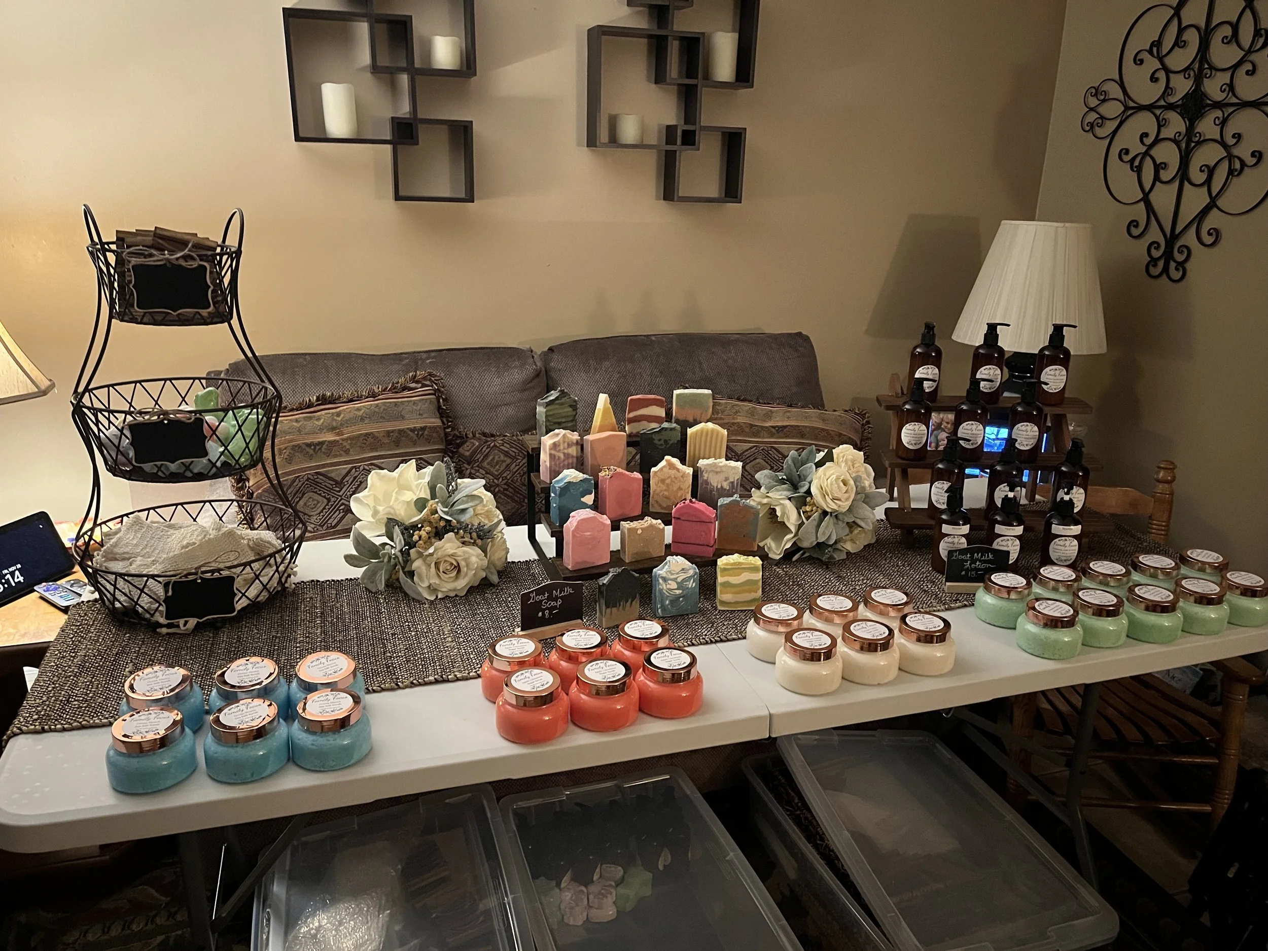 Display of homemade soaps, lotions, and other body care products on a table, with decorative flowers and signs labeling the products.
