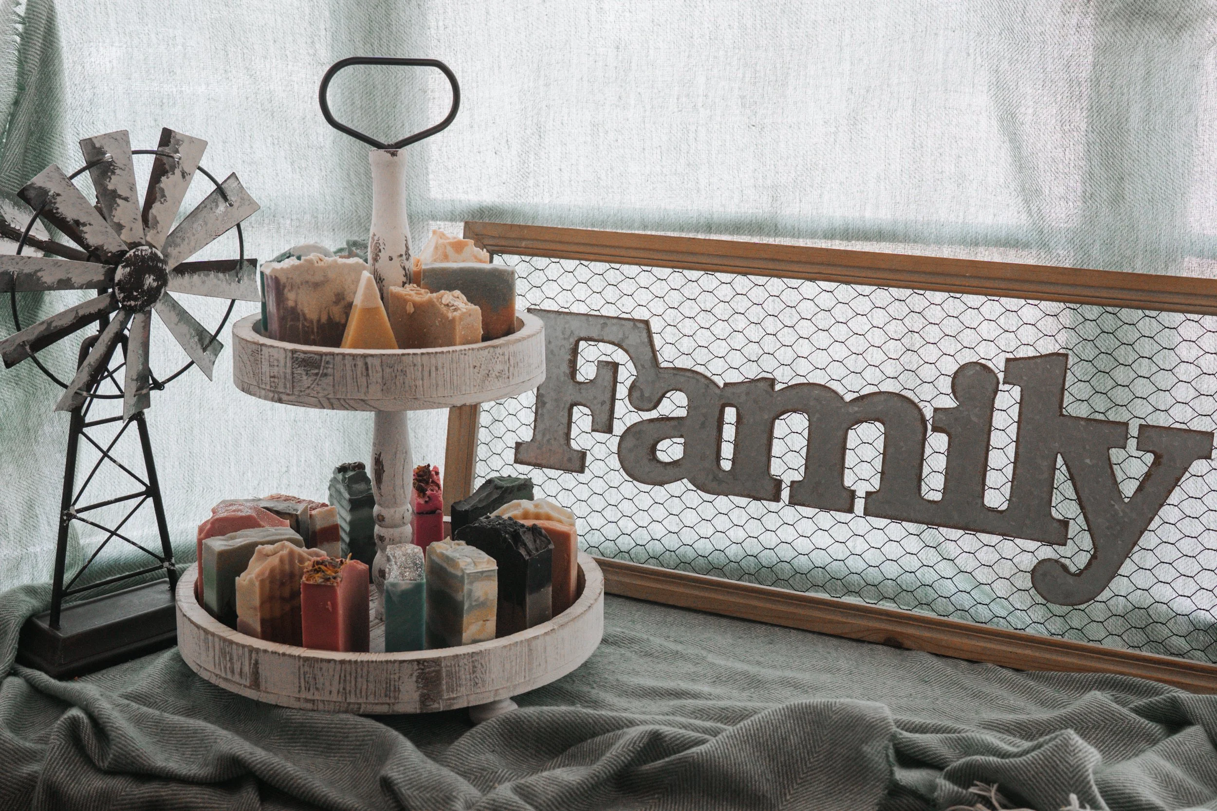 Display of decorative soap bars on a two-tiered white wooden stand, a metal windmill decoration, and a wooden framed 'family' sign with wire mesh backing, all set on a green cloth background.