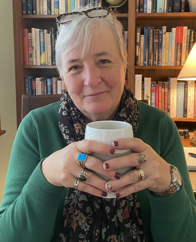 Author Julie Quinn shown with short gray hair, wearing a green sweater and decorative scarf, glasses on her head, holding a white mug in ring-bedecked hands, sitting in a room filled with books and a globe, with a lamp on a desk nearby.