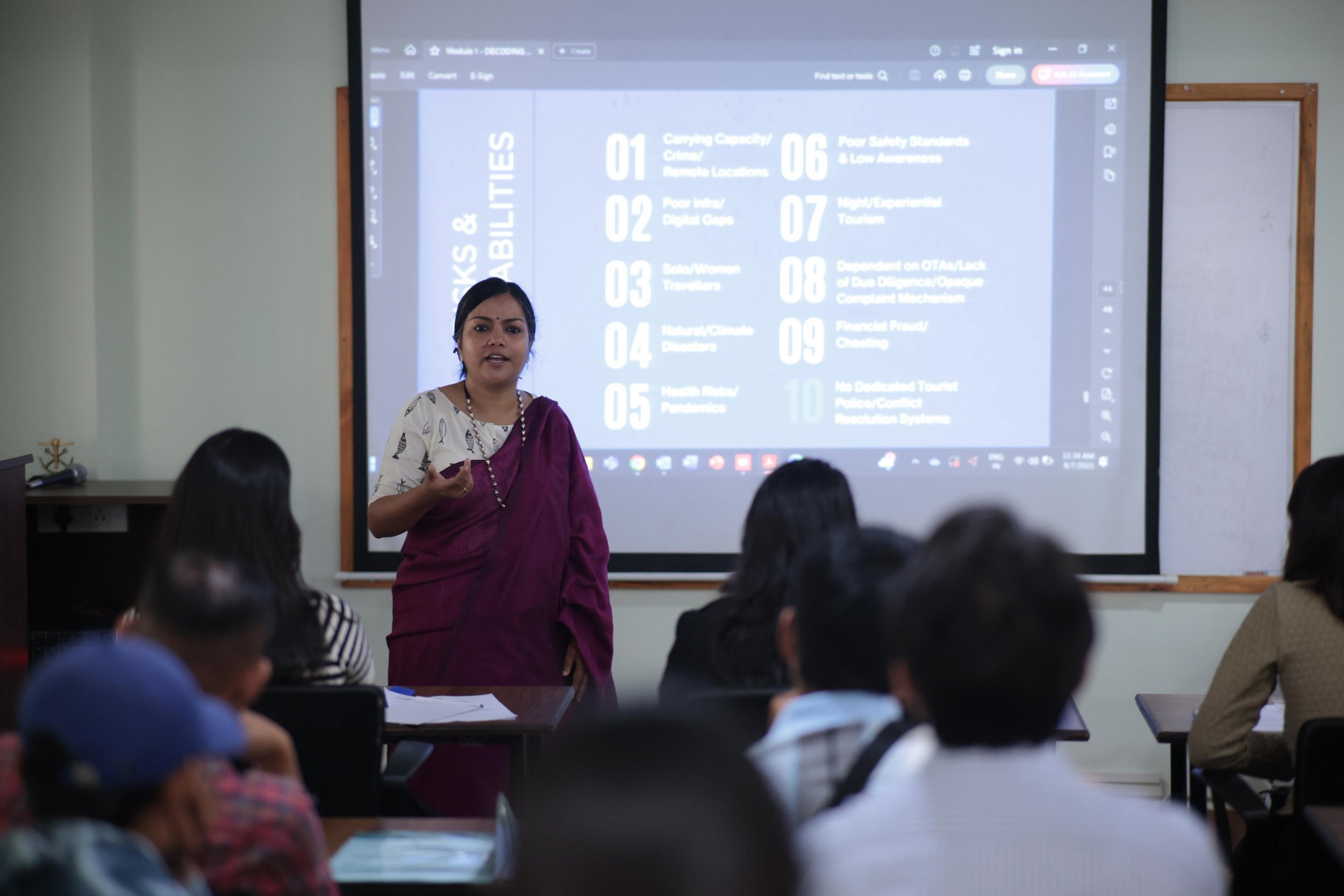 A woman wearing a traditional saree giving a presentation on sustainable tourism, with a slide titled "Risks & Vulnerabilities" projected on a screen behind her, listing ten points related to risks and vulnerabilities.