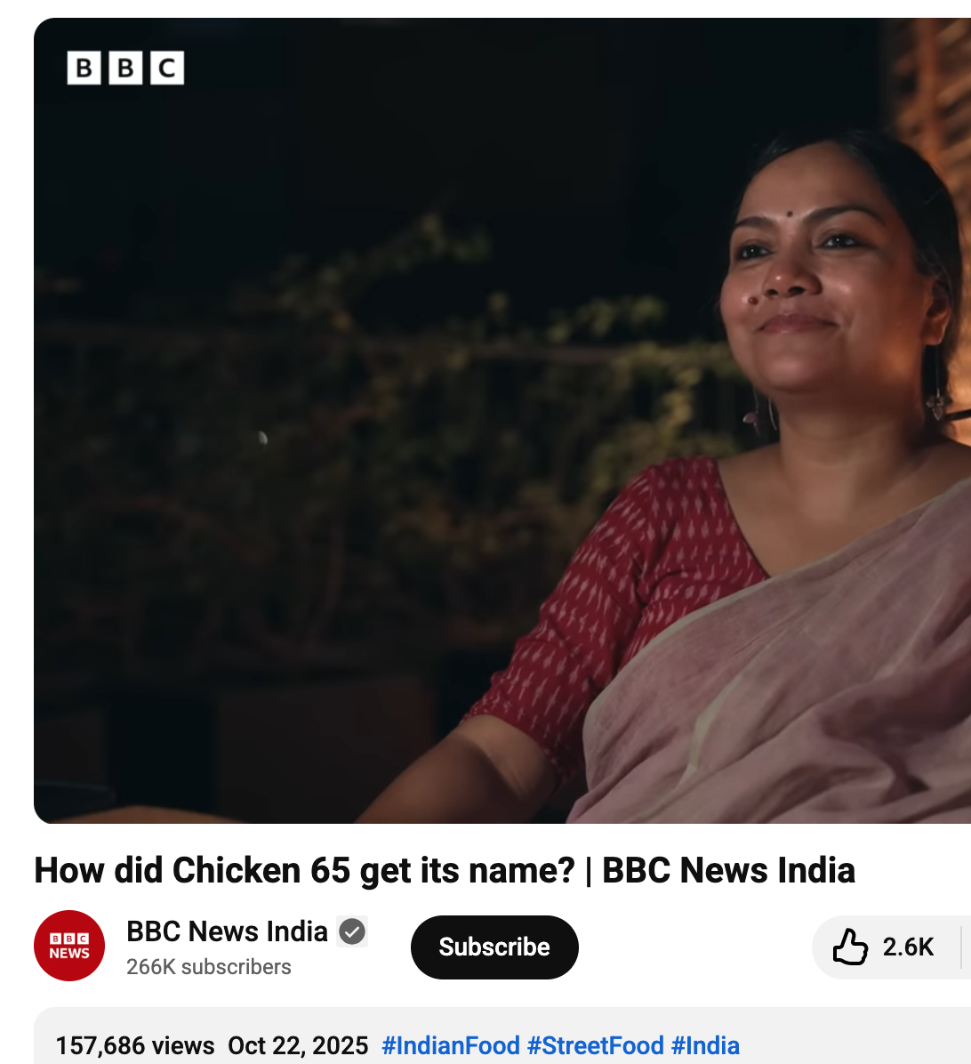 A woman smiling with a dark outdoor background, wearing a red and beige traditional Indian saree, during nighttime.