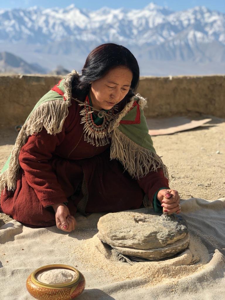 An elderly woman dressed in traditional clothing, kneeling on sand and grinding a stone with a smaller stone, with snow-capped mountains in the background.