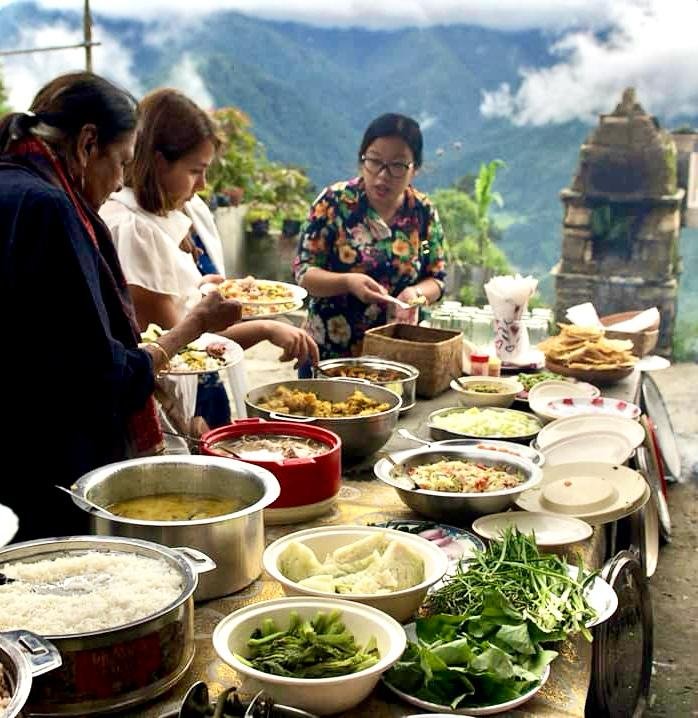 People serving themselves food from buffet spread outdoors with mountains in background.