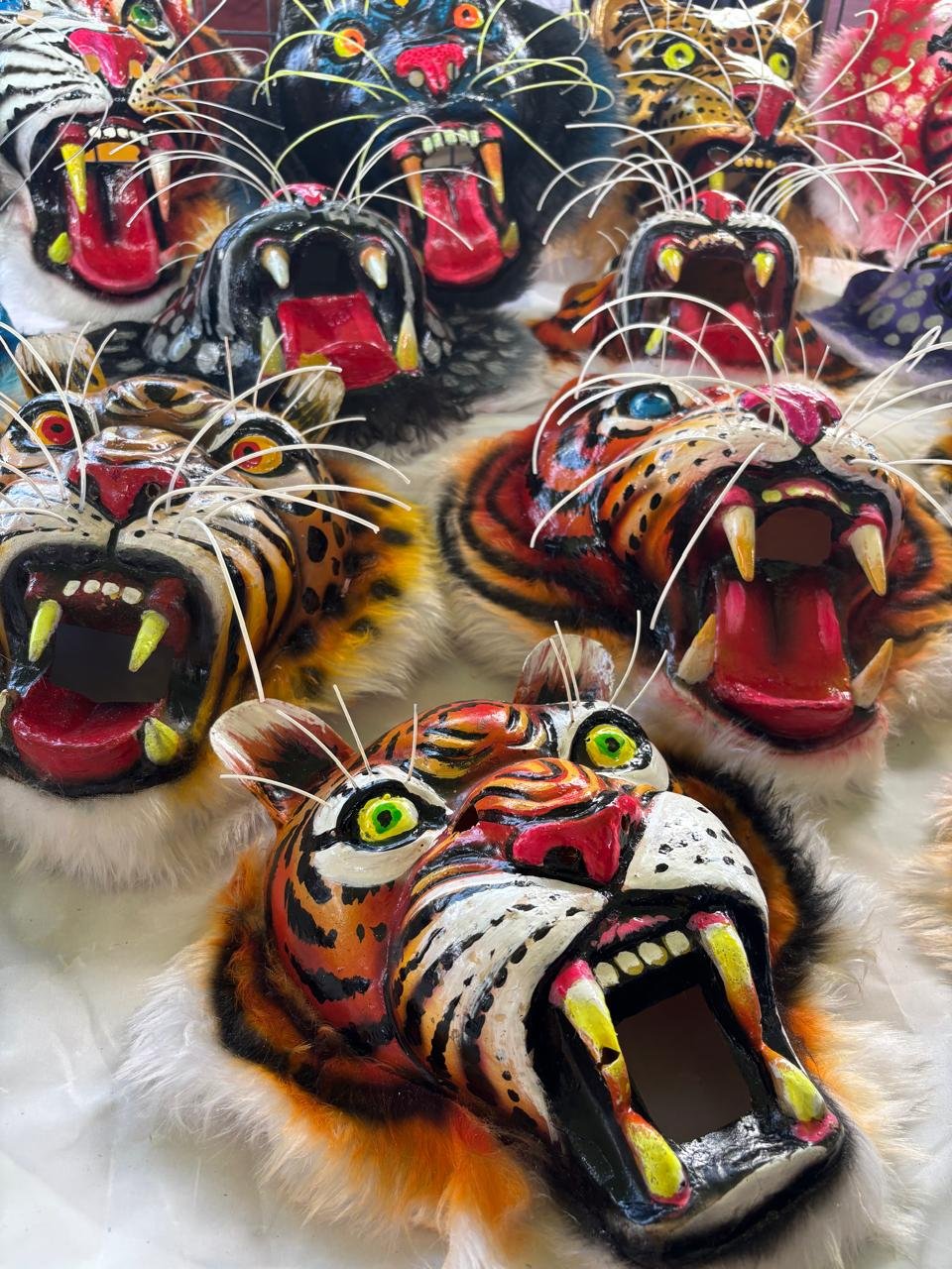 Colorful tiger and panther masks with open mouths exposing teeth, some with whiskers, in bright vivid colors and patterns, arranged together.