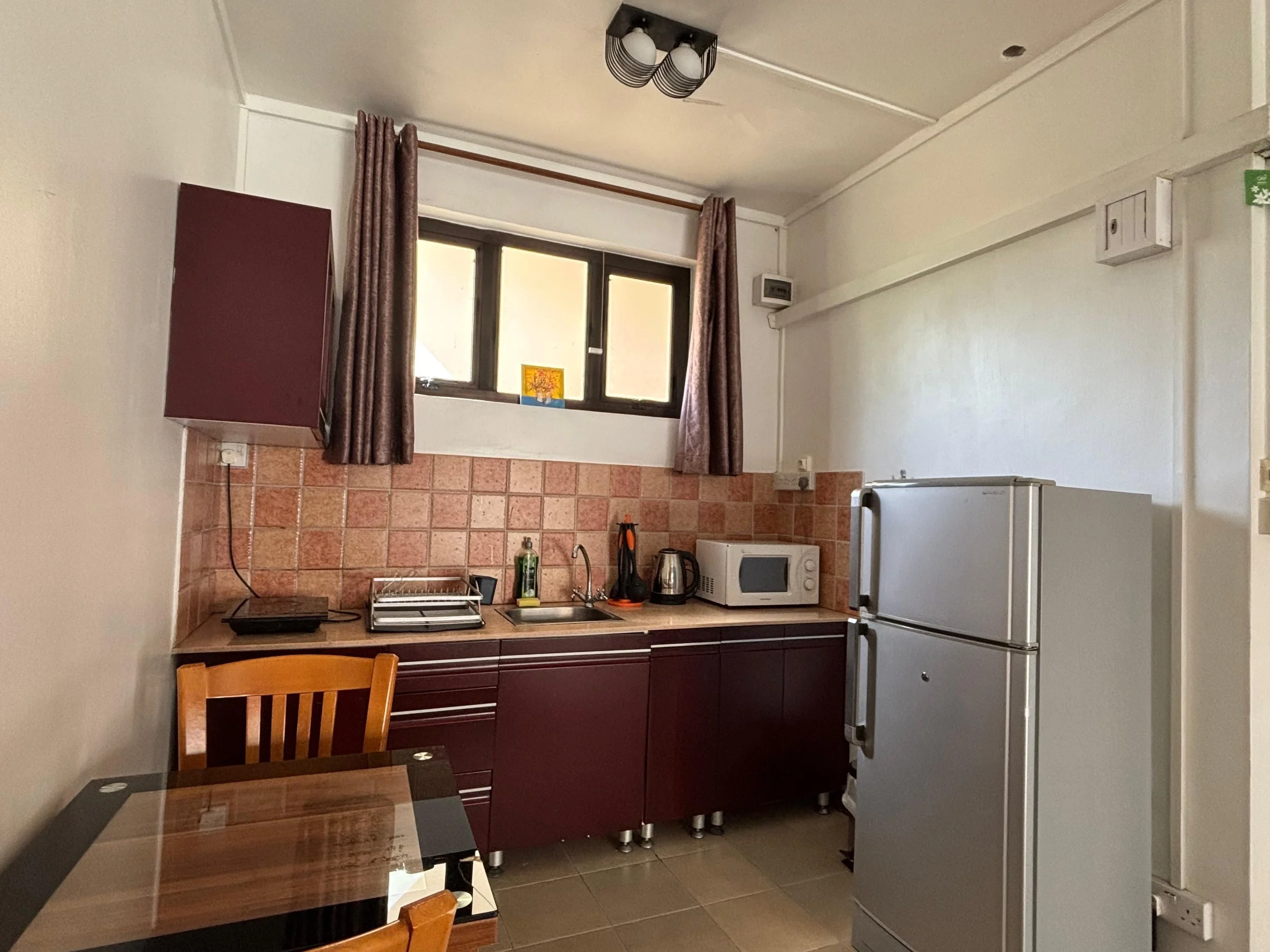 A kitchen with a cream wall, brown tile backsplash, dark wood cabinets, a window with pink patterned curtains, a small painting on the window sill, a microwave, a refrigerator, a kettle, and a brown dining chair.