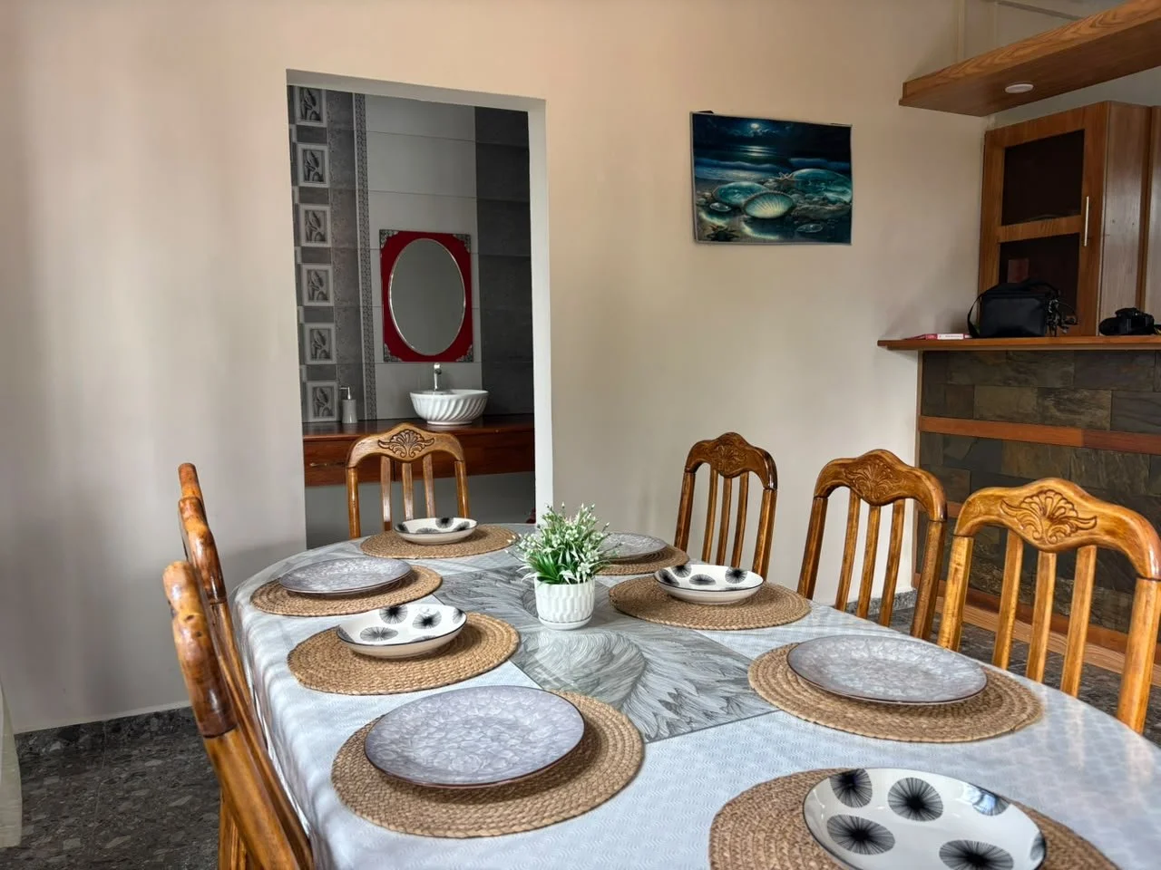 Dining table set with six placemats and matching plates, and a small flower centerpiece, in a room with a view of a bathroom through an open doorway.