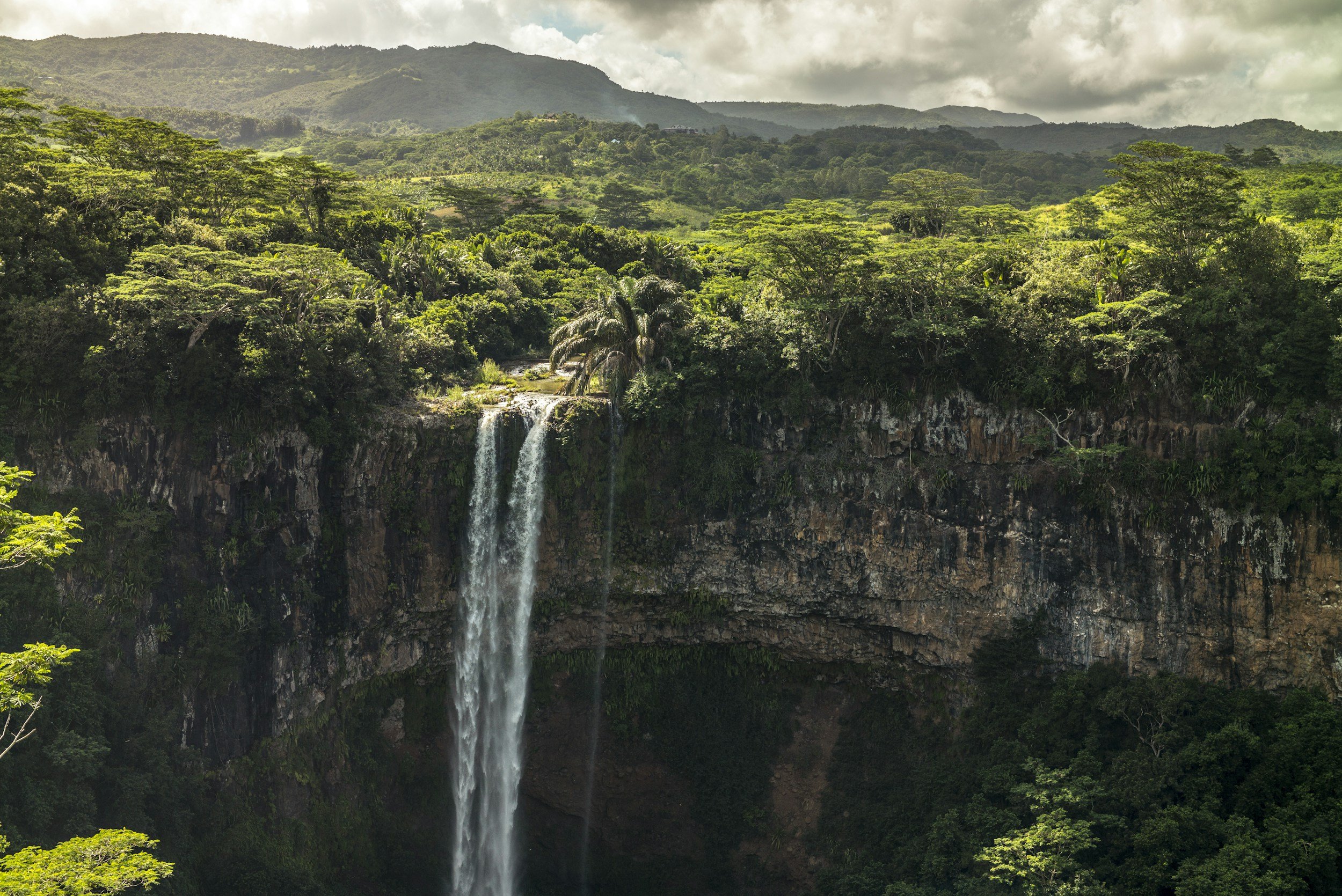 Lush green tropical forest with a waterfall cascading down a rocky cliff, mountain range in the background, cloudy sky overhead.