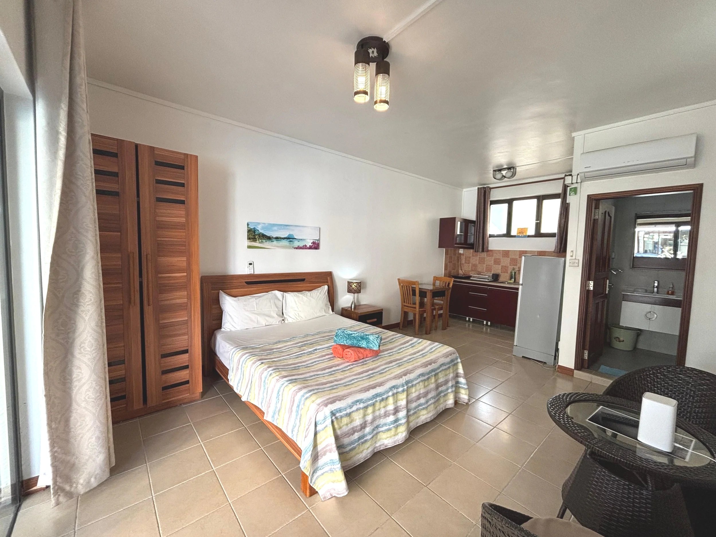 Interior of a bedroom with a bed, a wooden wardrobe, a small table with a lamp, a kitchenette with cabinets, a window with curtains, a door, and a wicker chair with a small table.