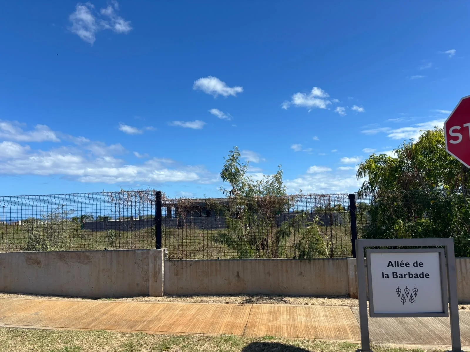 A fenced outdoor area with a sign that reads 'Allée de la Barade,' a stop sign on the right, and blue sky with clouds overhead. Green trees and dry vegetation are visible behind the fence.