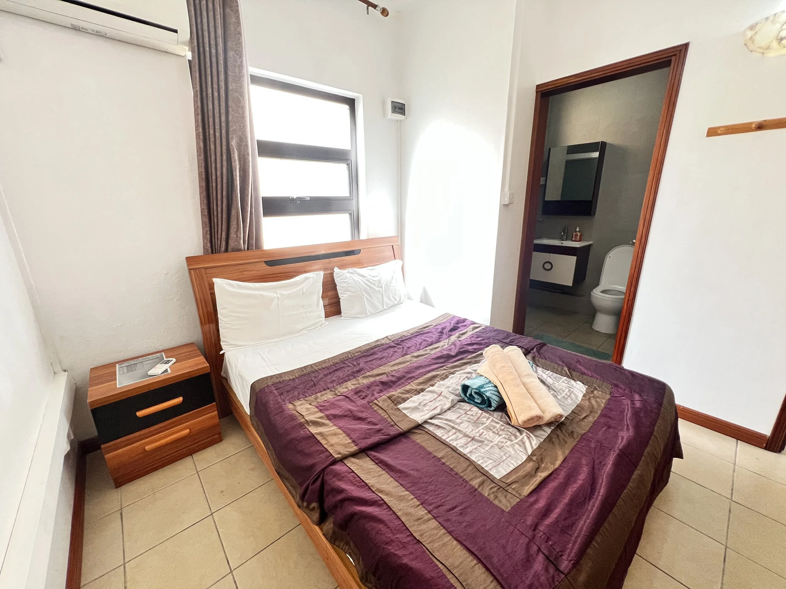 A hotel room with a bed covered in purple and beige striped bedding, two white pillows, and several folded towels on top. There is a wooden nightstand with a remote control, an air conditioning unit above the window, and a doorway leading to a small bathroom with a toilet, sink, and wall-mounted mirror.