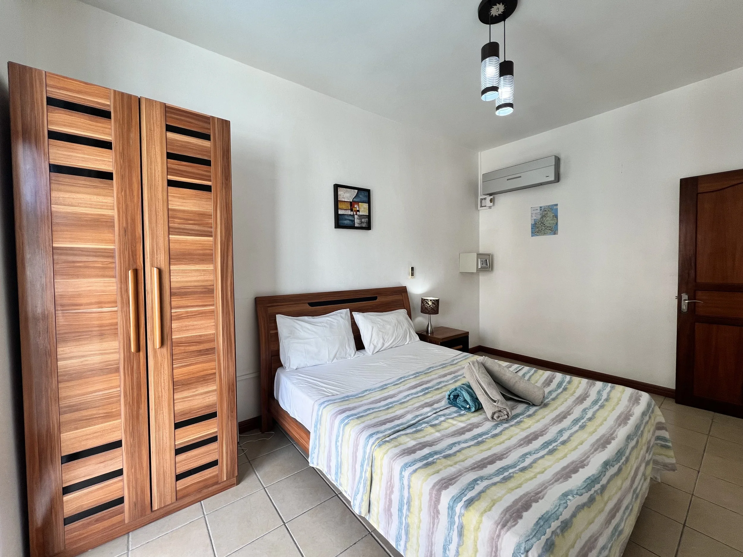 A bedroom with a bed, wooden wardrobe, small bedside table with a lamp, and wall decorations, including a small framed painting and a map, in a cozy and minimalistic style.