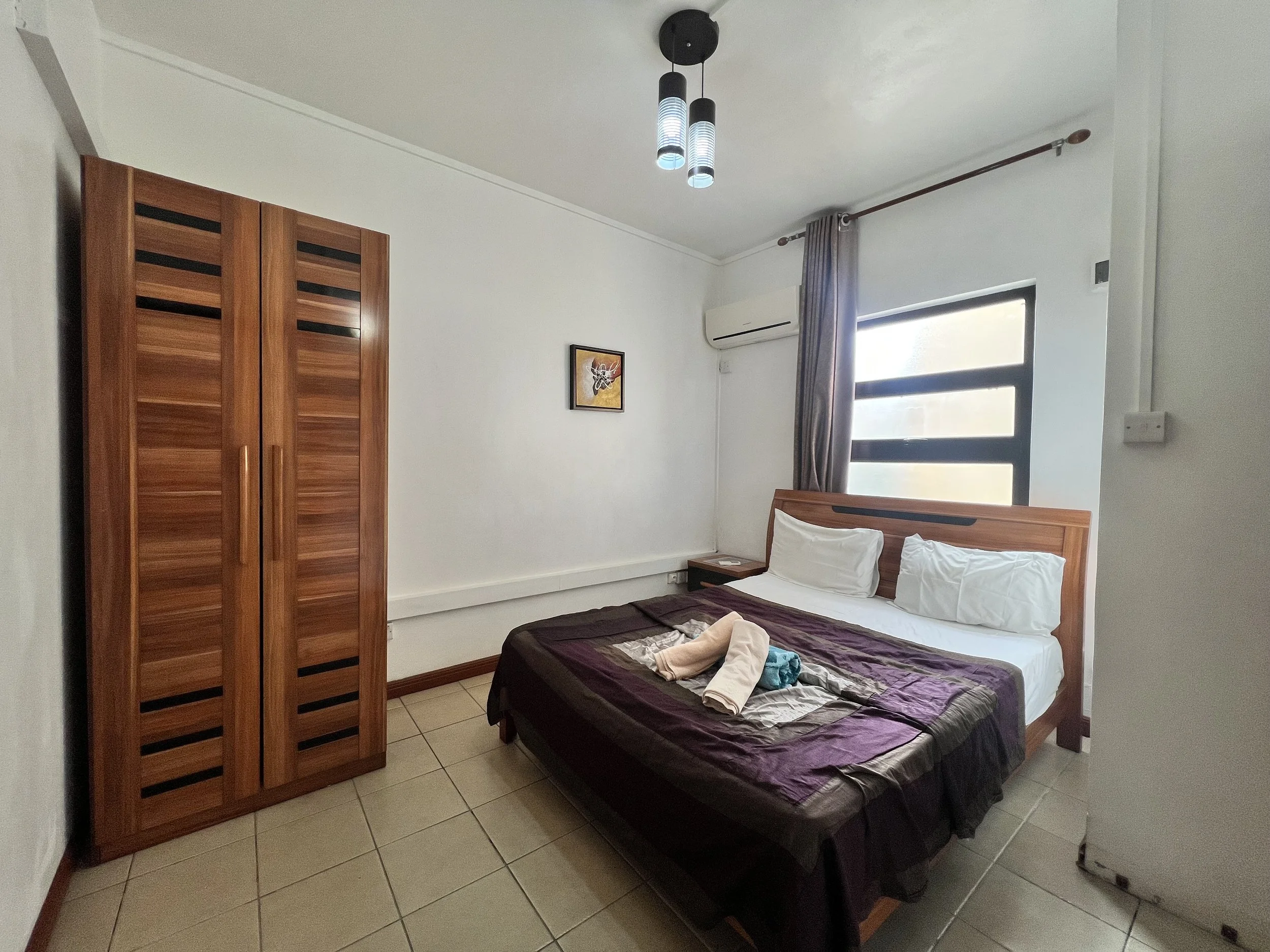 A bedroom with a bed, pillow, and blanket, a wooden wardrobe, a framed artwork on the wall, a window with curtains, a ceiling light fixture, and an air conditioner.