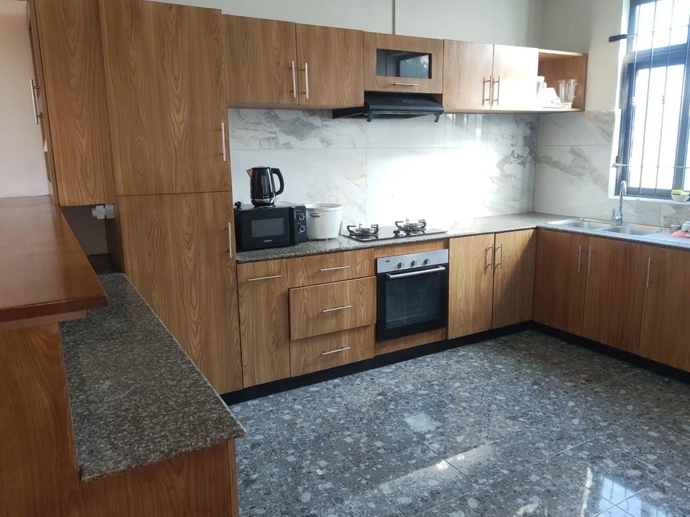 Kitchen with wooden cabinets, granite countertops, stovetop, oven, microwave, electric kettle, rice cooker, and a window above the sink.