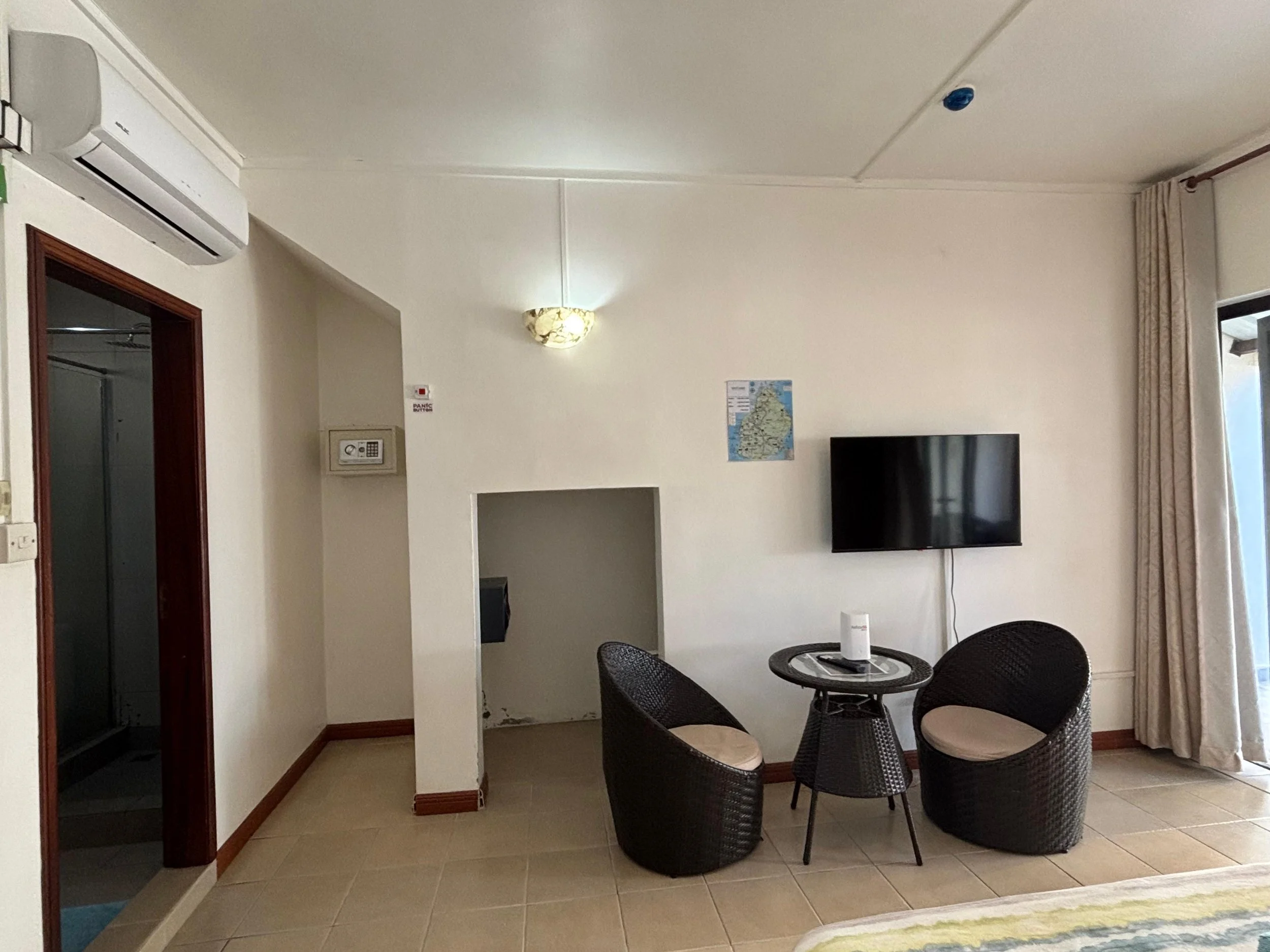 Living room with white walls, a wall-mounted TV, a small dining table with four wicker chairs, a wall light, an air conditioning unit, a map, a fire alarm, and curtains by a window.